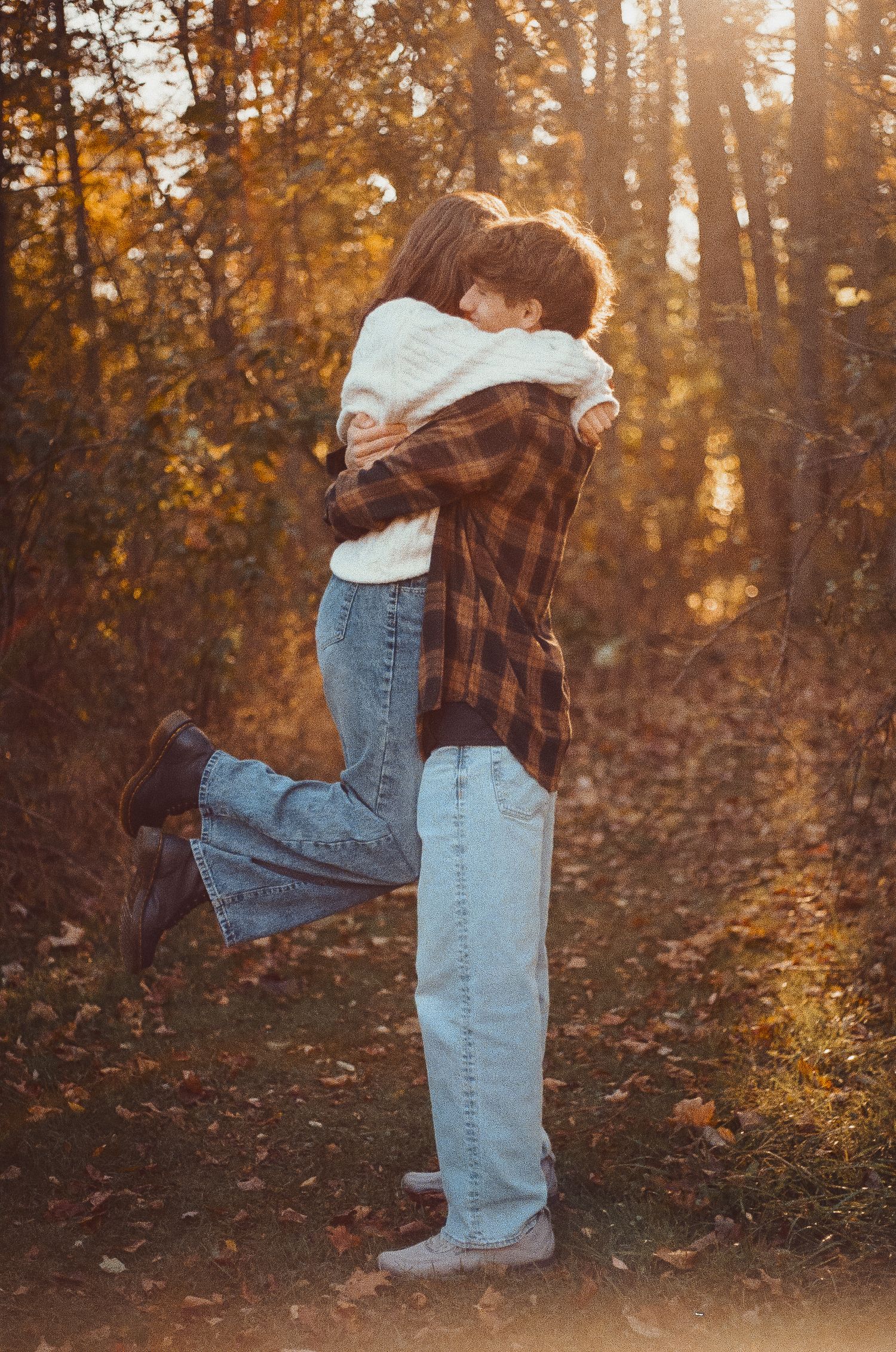 A couple in plaid and denim embrace in a romantic lift pose during golden hour in an autumn forest setting.