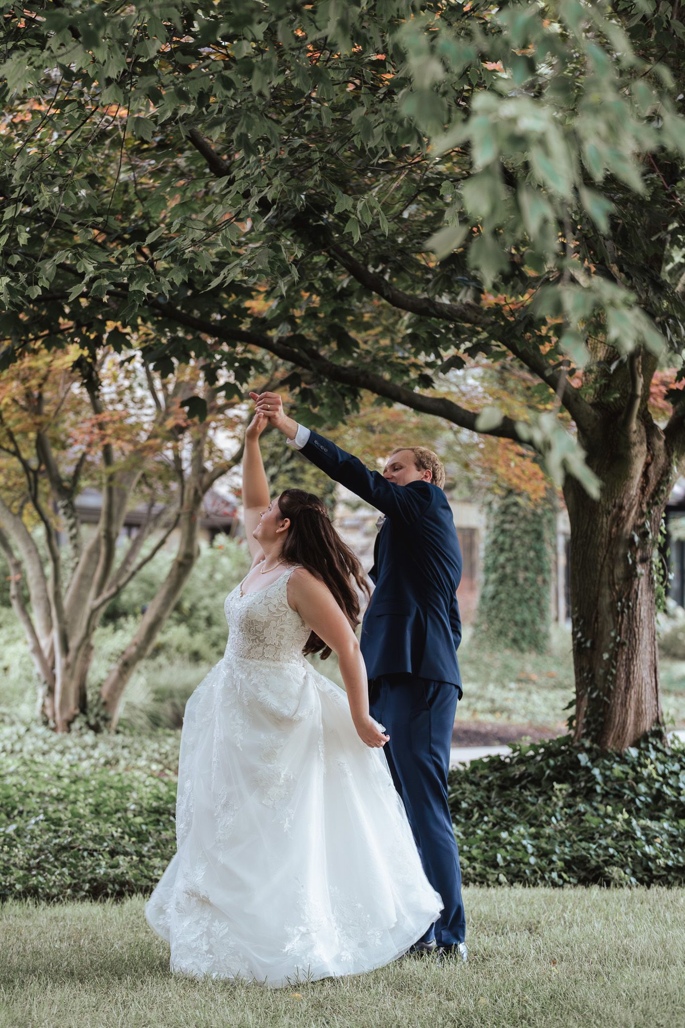 Couple dances playfully on grass under trees during their wedding photoshoot.