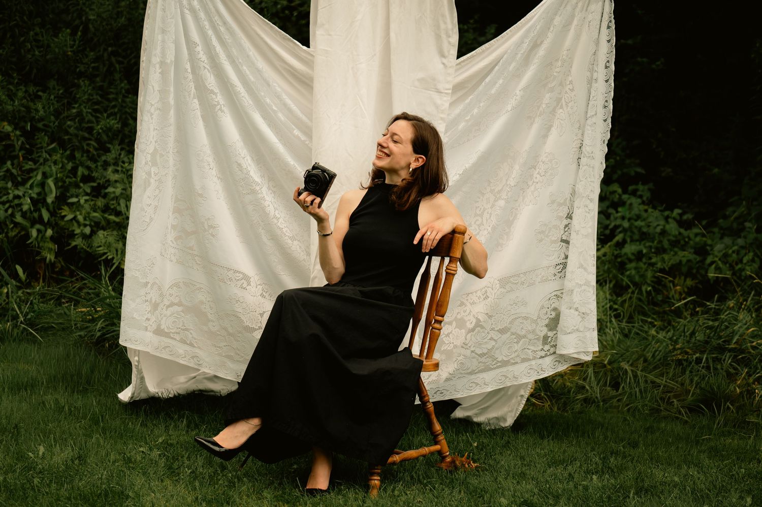 Woman in black dress sitting on wooden chair outdoors, laughing and holding small black object near white lace backdrop.
