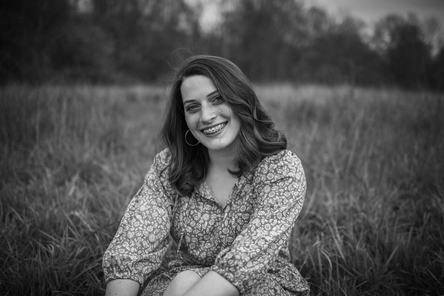 Candid portrait of someone in a floral print dress sitting in a grassy field on a cloudy day.