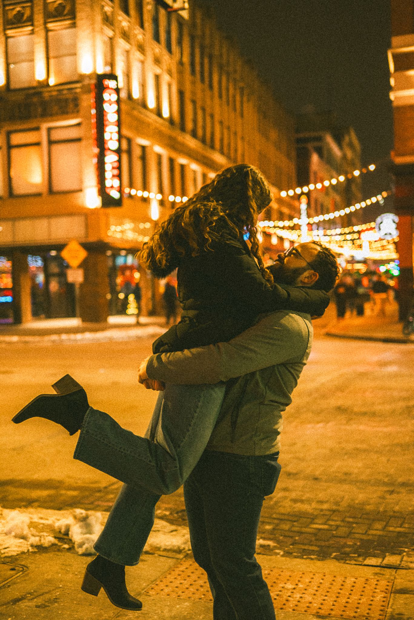 Romantic couple dancing and embracing on illuminated city street at night with string lights glowing overhead.