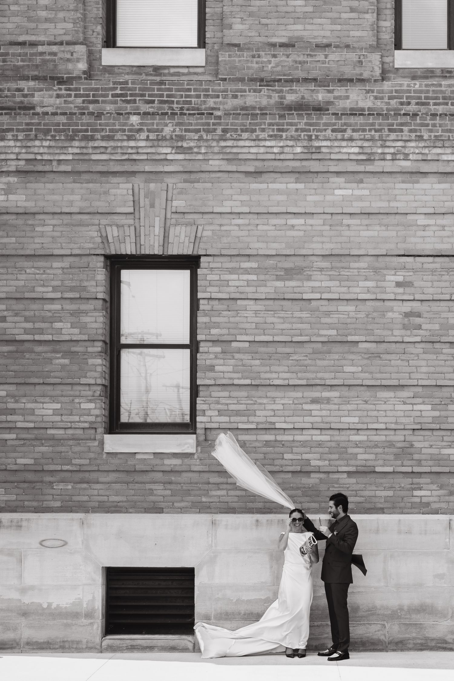 Black and white photo of bride and groom standing against brick building, veil flowing in wind.