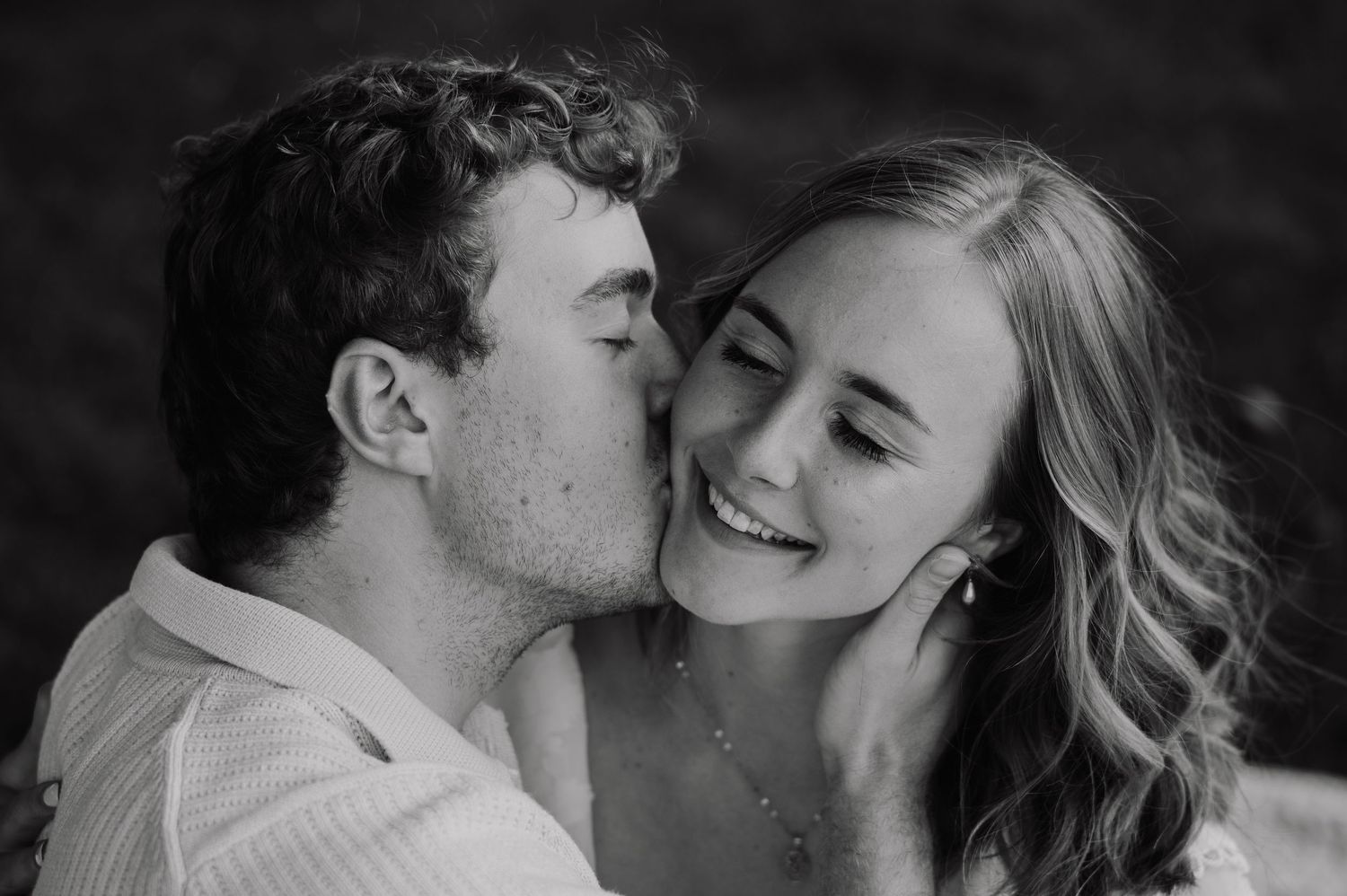 Black and white photo of a young couple, man kissing womans cheek as she smiles warmly.
