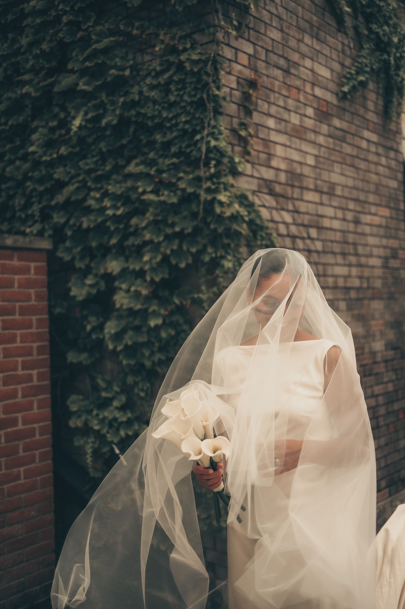 Bride in white gown and flowing veil holds calla lily bouquet beside ivy-covered brick wall.