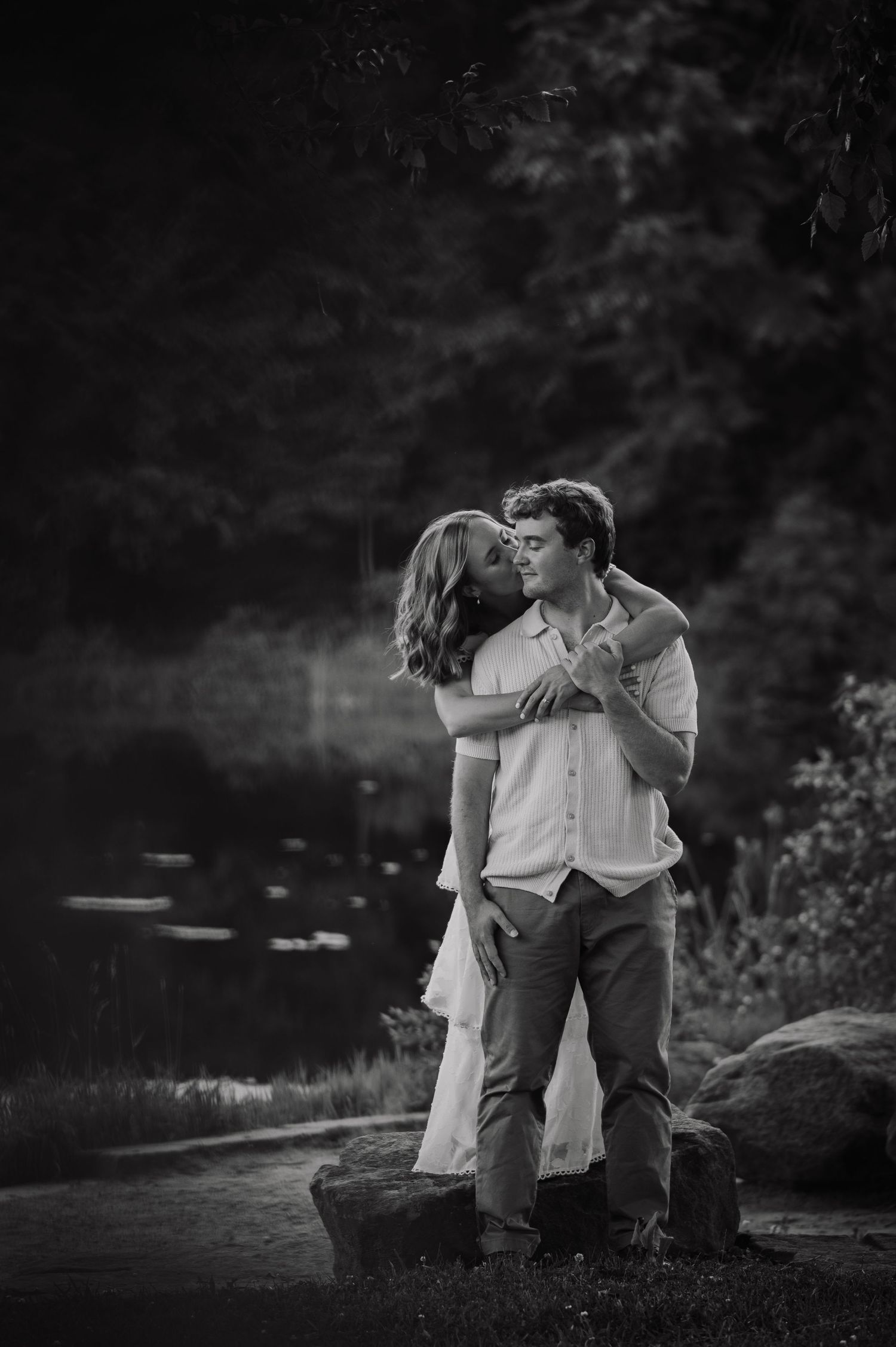 Black and white photo of a couple sharing a romantic kiss outdoors near a pond surrounded by lush trees.