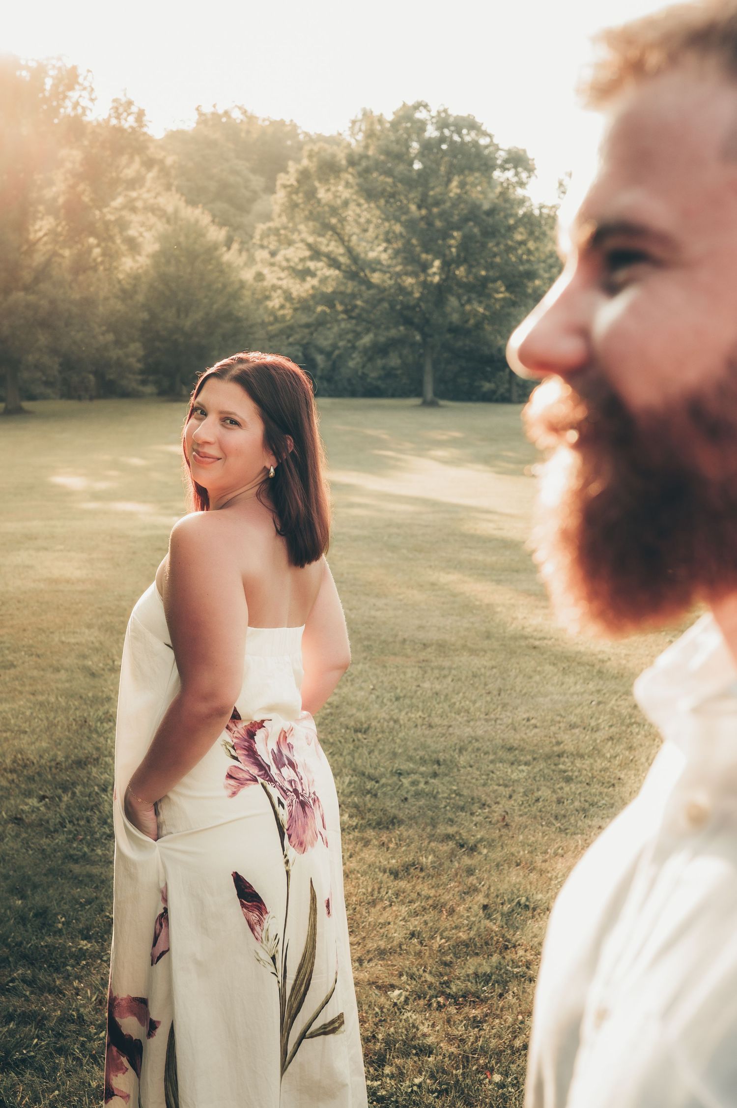 A woman in a floral white dress smiles over her shoulder at a bearded man in a sunlit park.