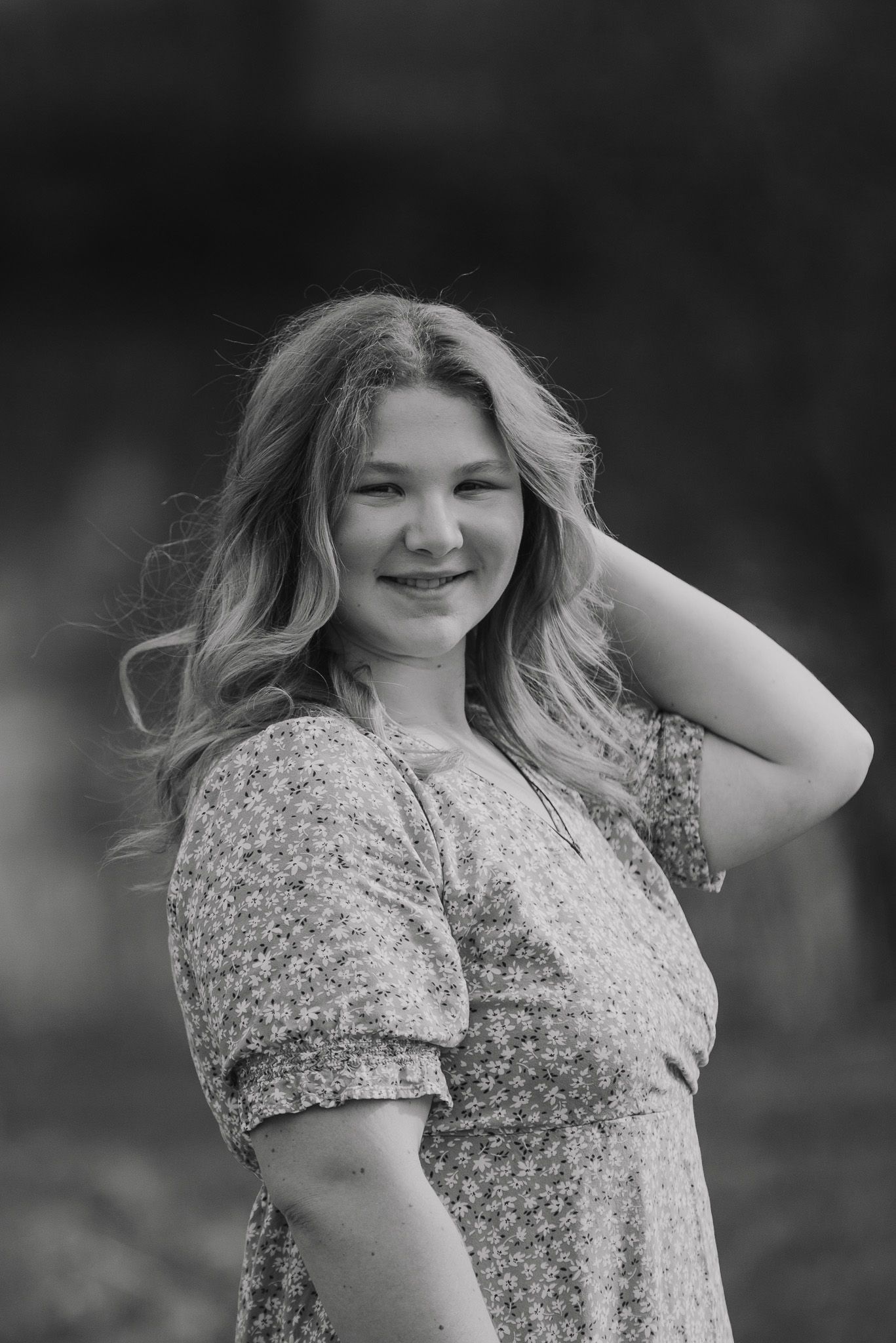 Black and white portrait of someone in a floral dress touching their hair outdoors.