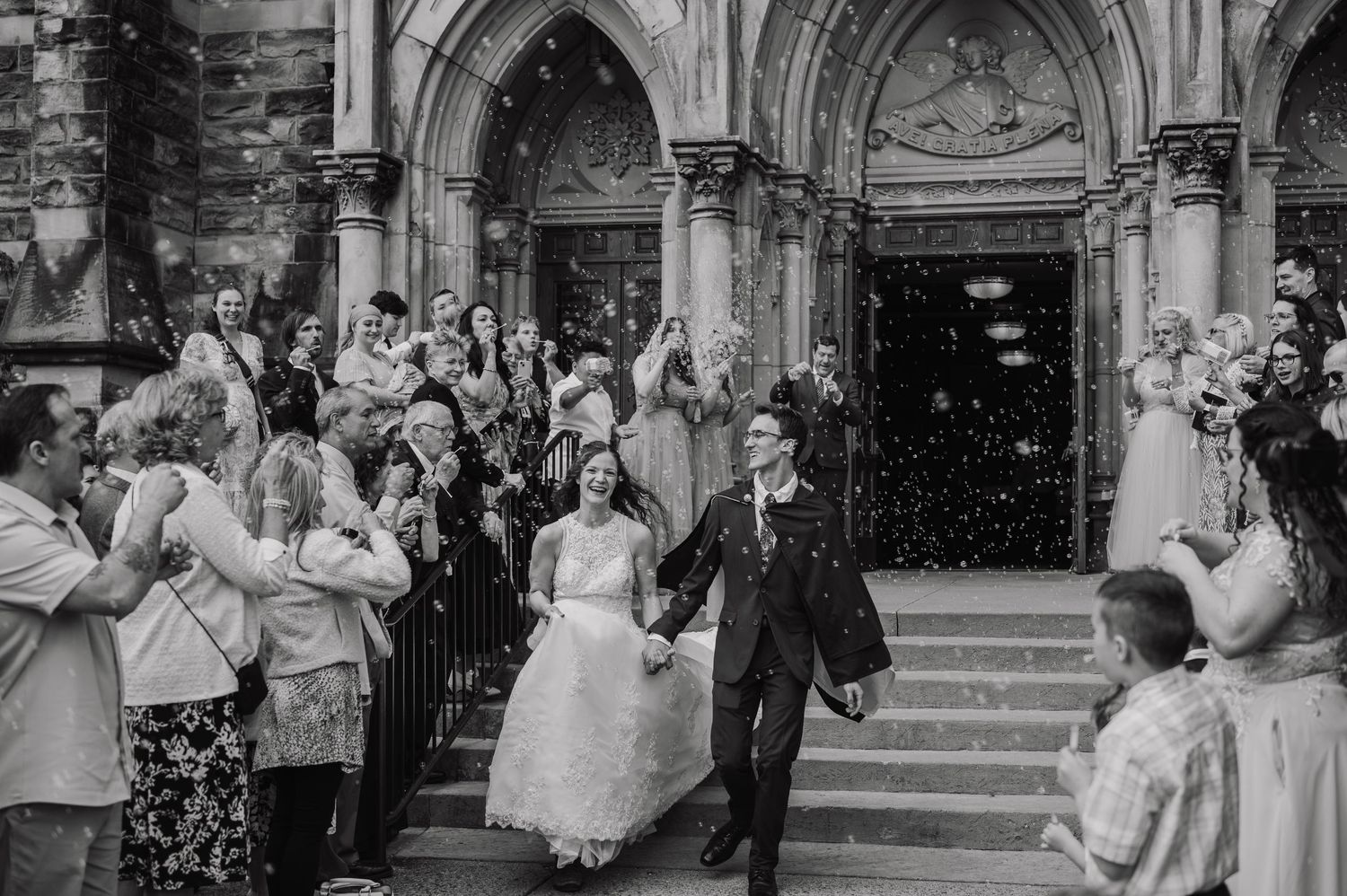 Black and white photo of newlyweds exiting church amid confetti, surrounded by cheering guests.