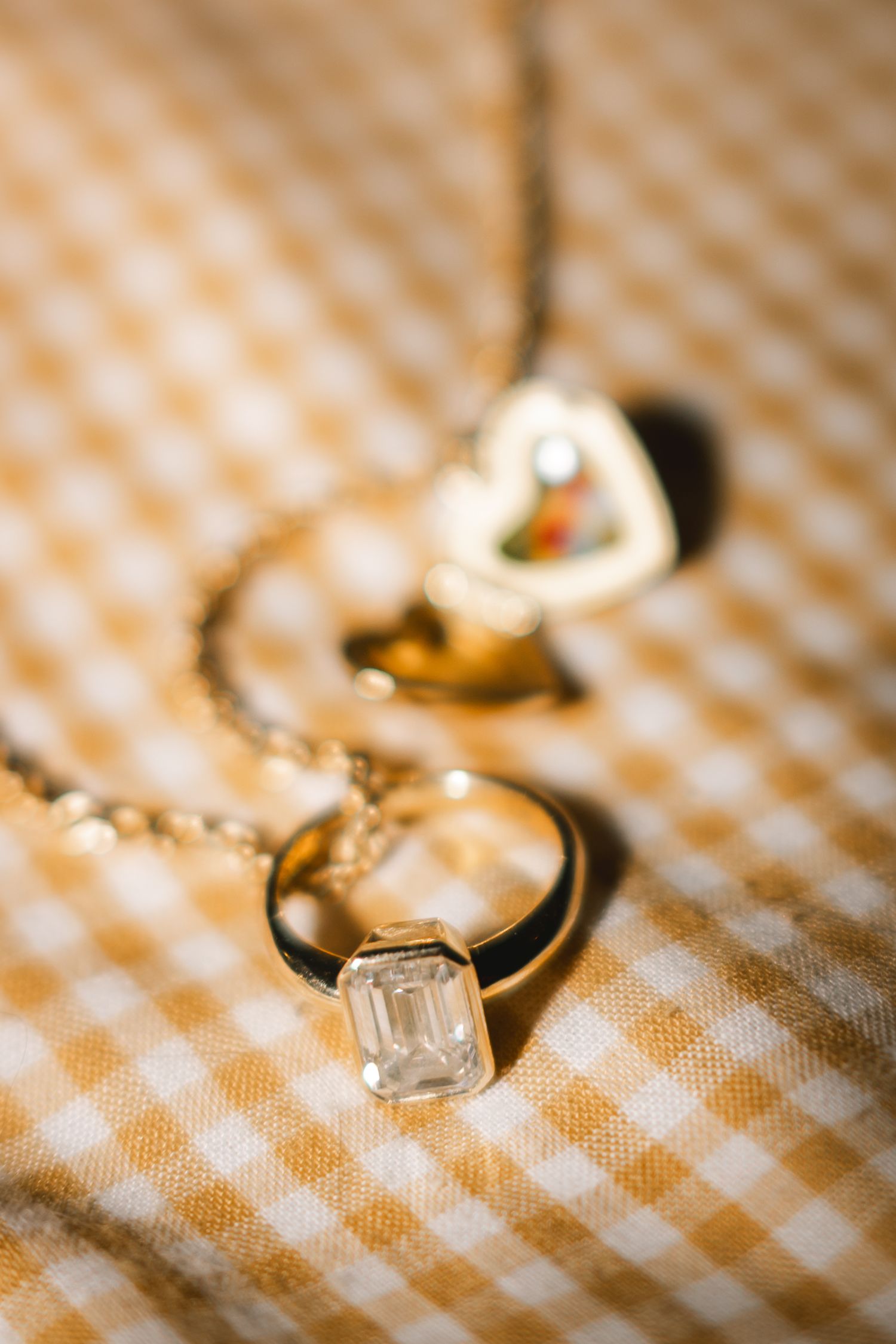 Gold ring with emerald-cut diamond pendant necklace on yellow gingham fabric with heart locket in background.