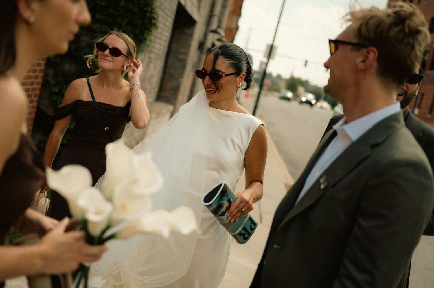 Bride in white dress with veil holds teal clutch, walks with wedding party in sunglasses on urban street.