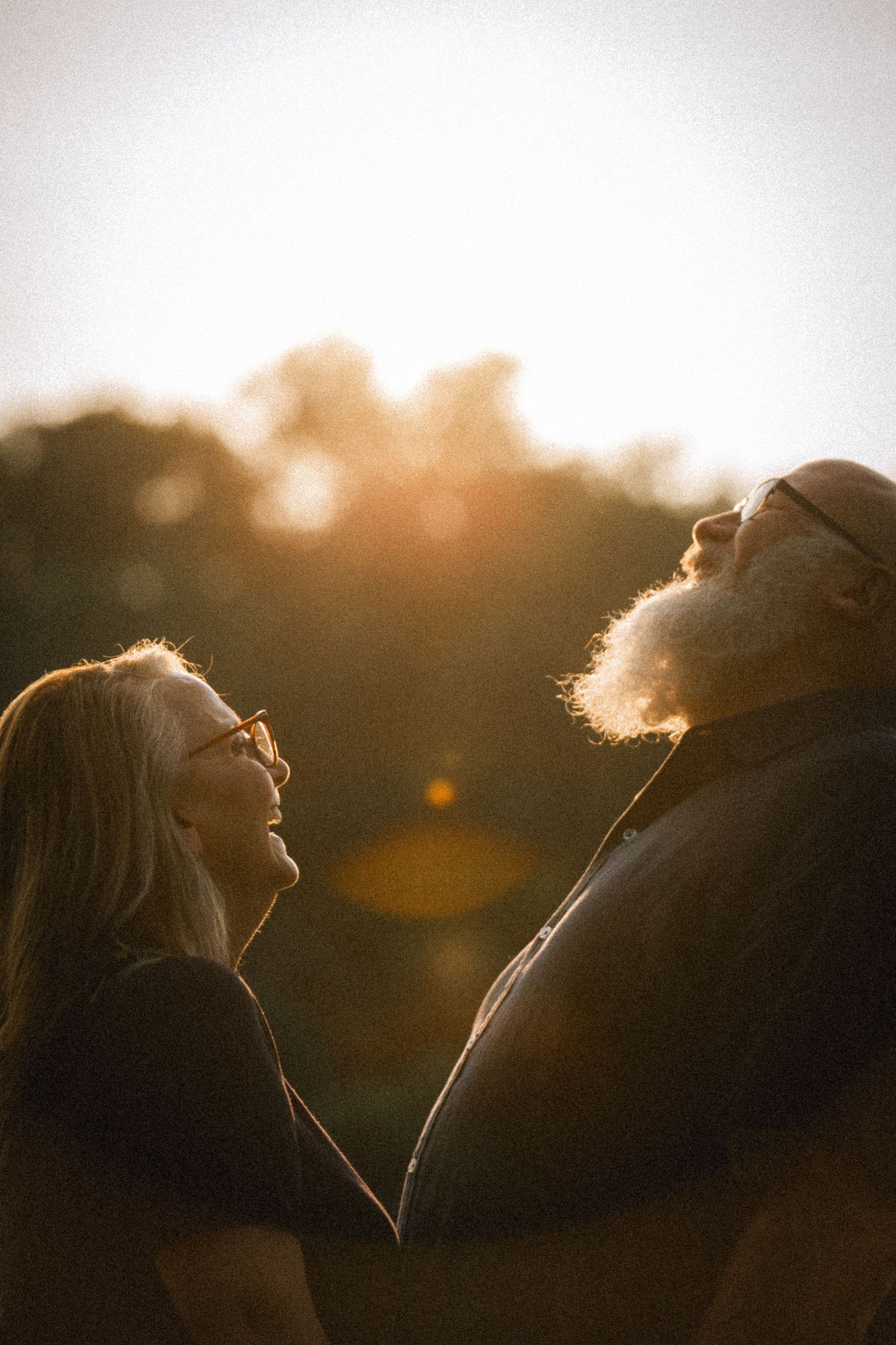 Elderly couple sharing a tender moment at golden hour sunset, woman with glasses and man with white beard looking up together.