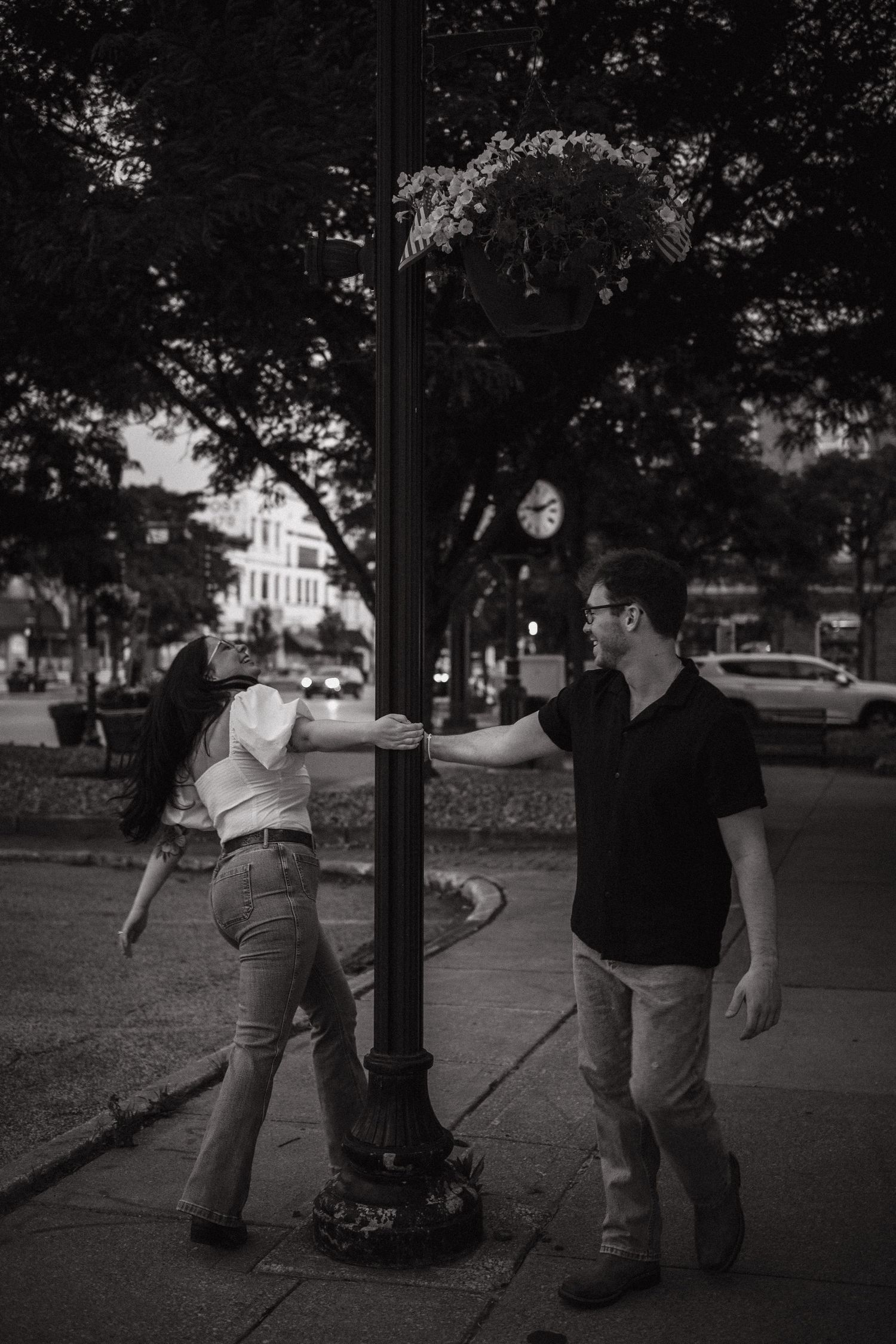 Black and white photo of a couple playfully posing around a street lamp on a city sidewalk at dusk.