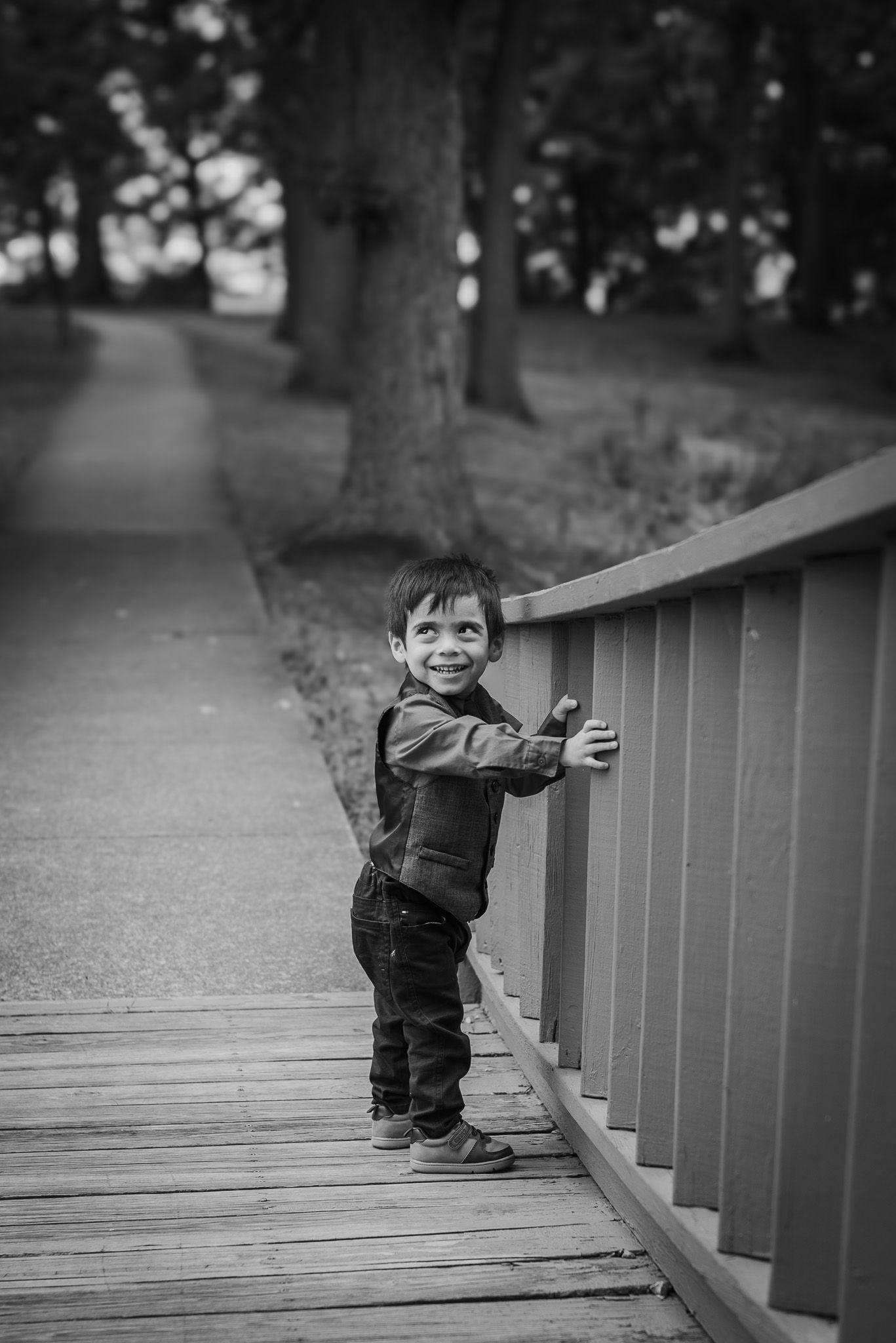 A small child in denim explores a wooden boardwalk railing in black and white.