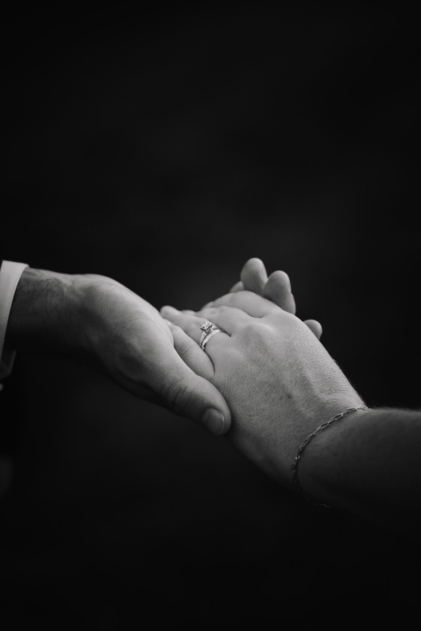 Black and white photo of two hands holding, one wearing a diamond engagement ring and delicate bracelet.