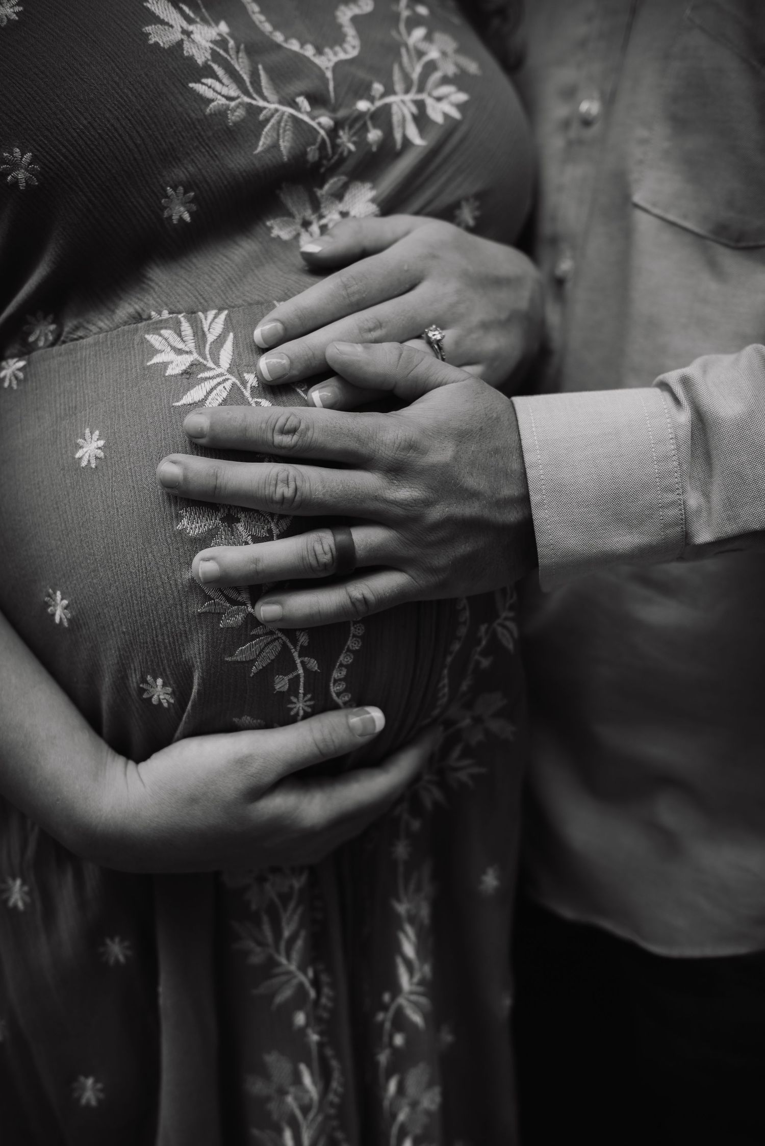 Black and white maternity photo of couple cradling pregnant belly, wearing wedding rings, in floral dress.