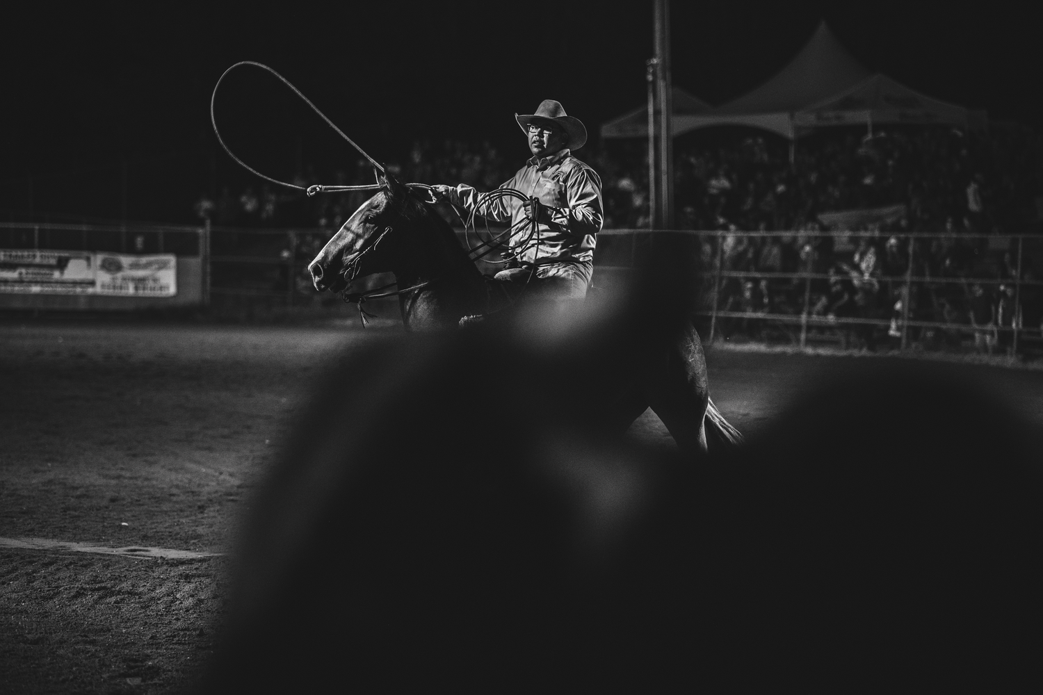 Black and white photo of cowboy on horseback swinging lasso at nighttime rodeo.