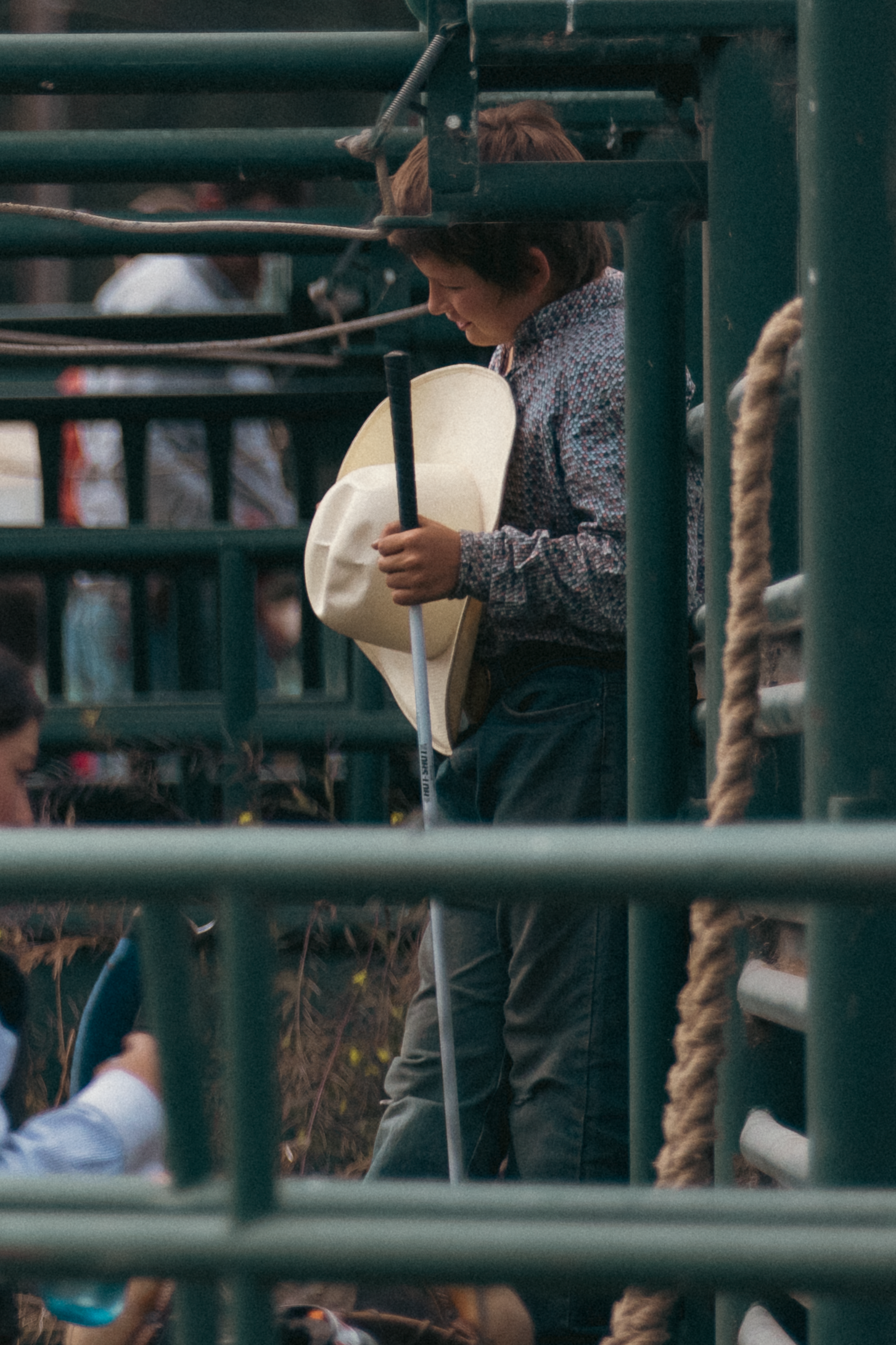 A young child holds a cowboy hat while standing near rodeo chutes, watching the event unfold.