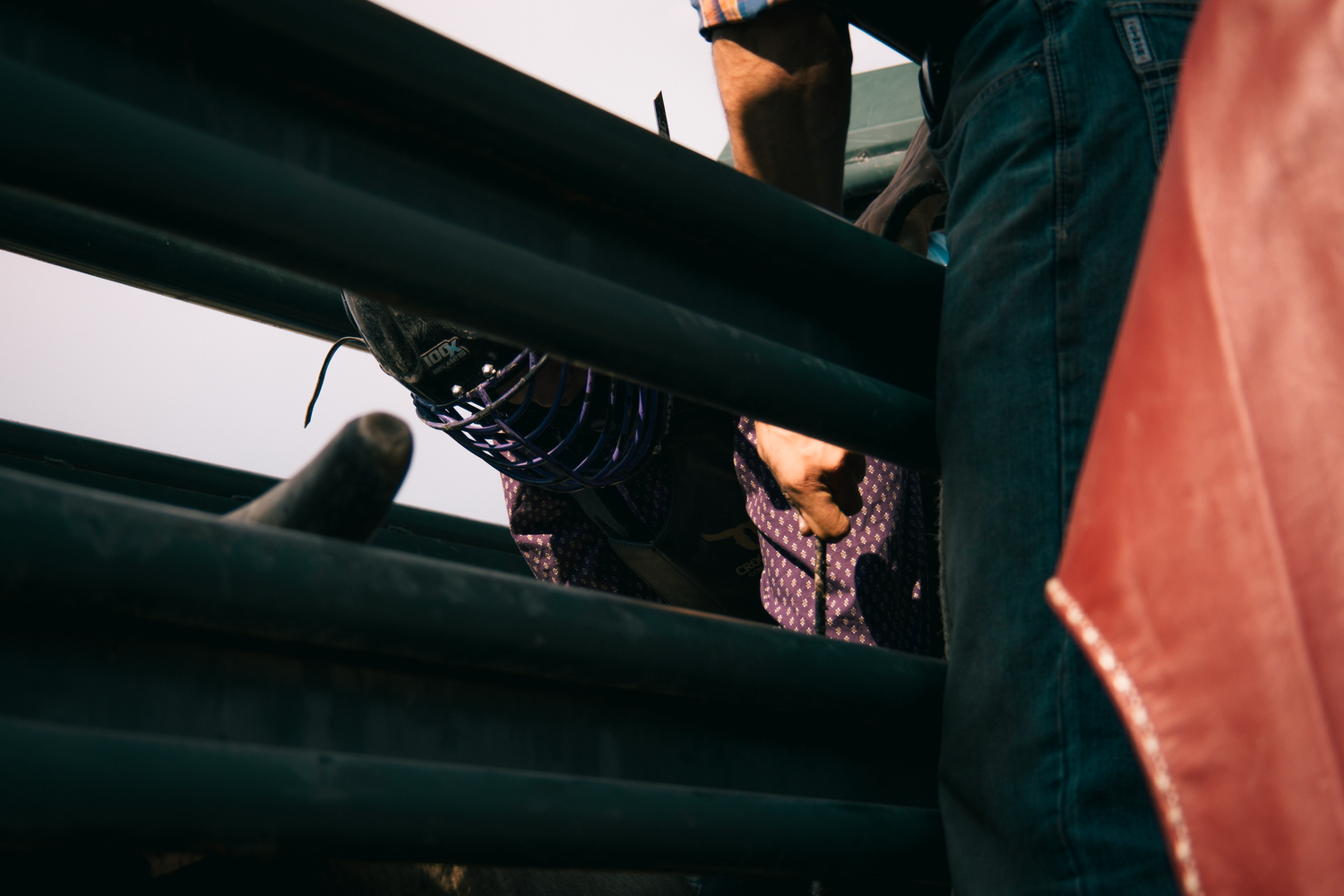Cowboy boots visible through green metal rodeo chute rails, with riders preparing for a bull riding event.