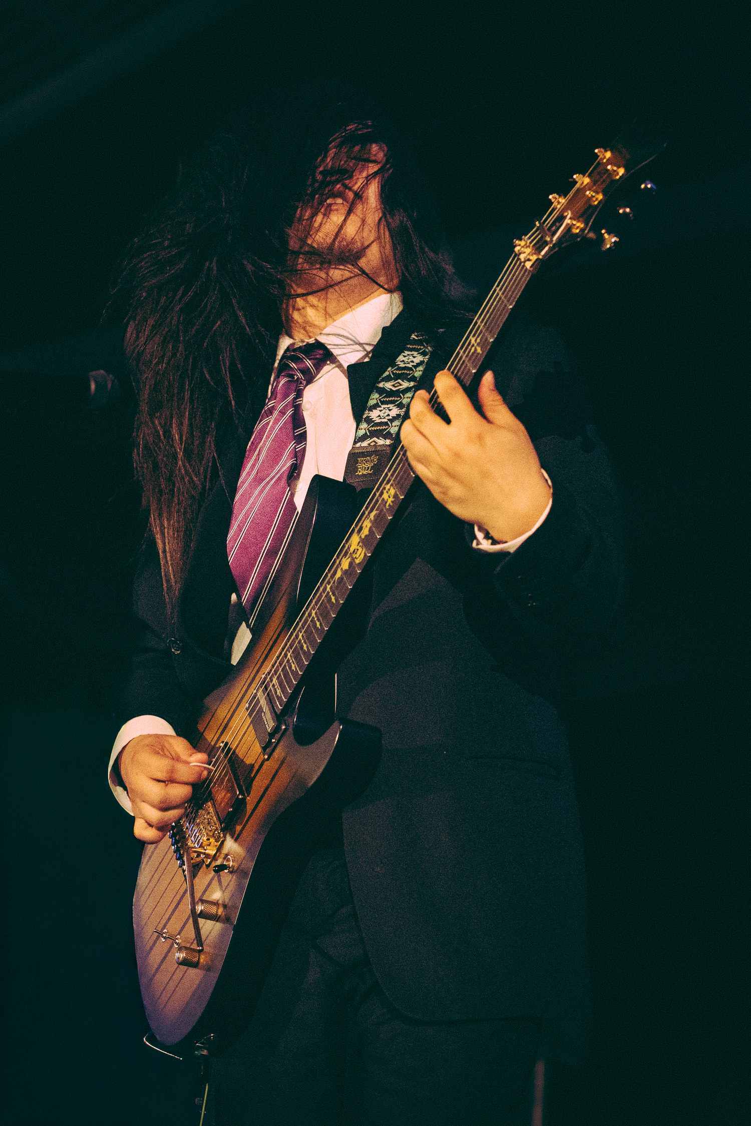Long-haired guitarist in suit and tie shredding electric guitar on dark concert stage with hair flying.