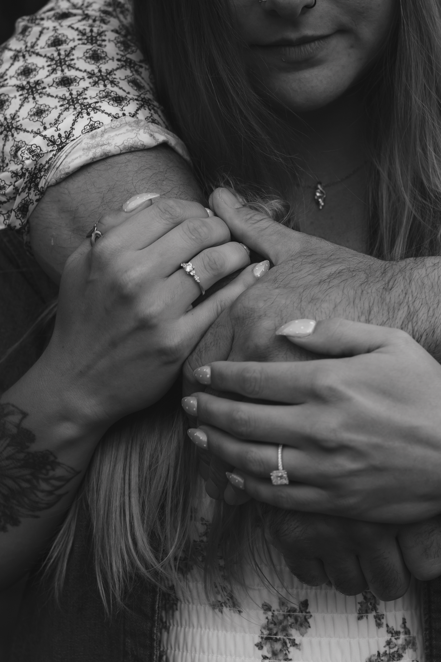 Black and white photo of a couple embracing, showcasing engagement rings on intertwined hands with a tattoo visible.