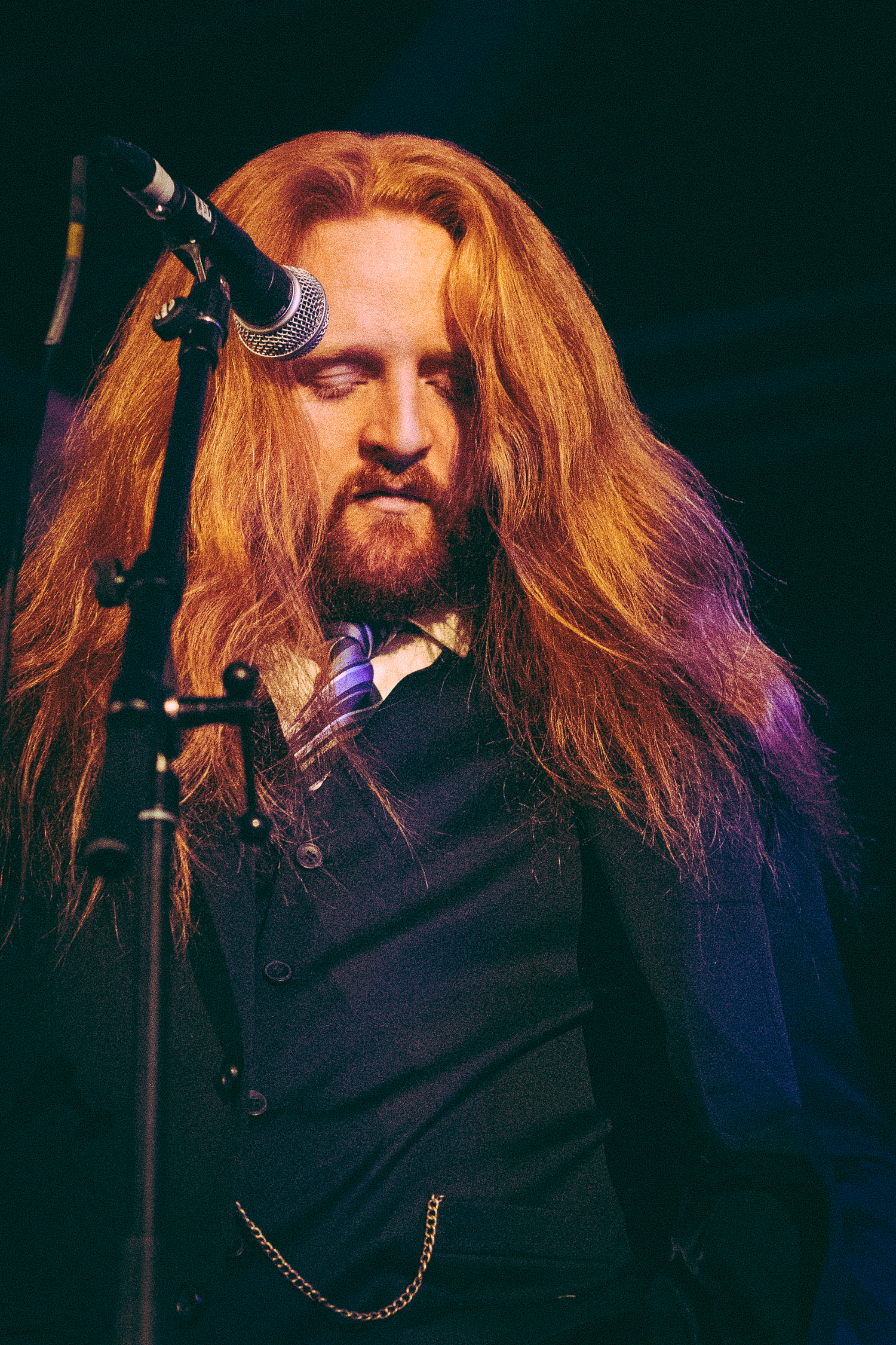 Long-haired redhead male vocalist performing on stage at a live music concert with microphone stand.