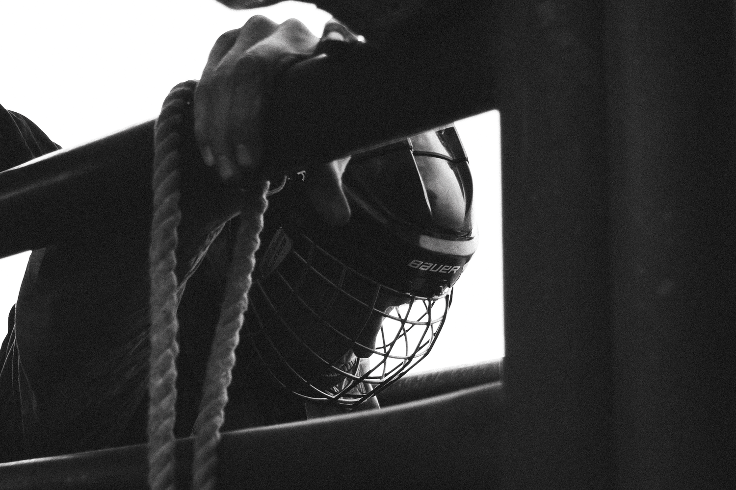 Black and white photo of a hockey player holding a helmet and rope near a rink board.
