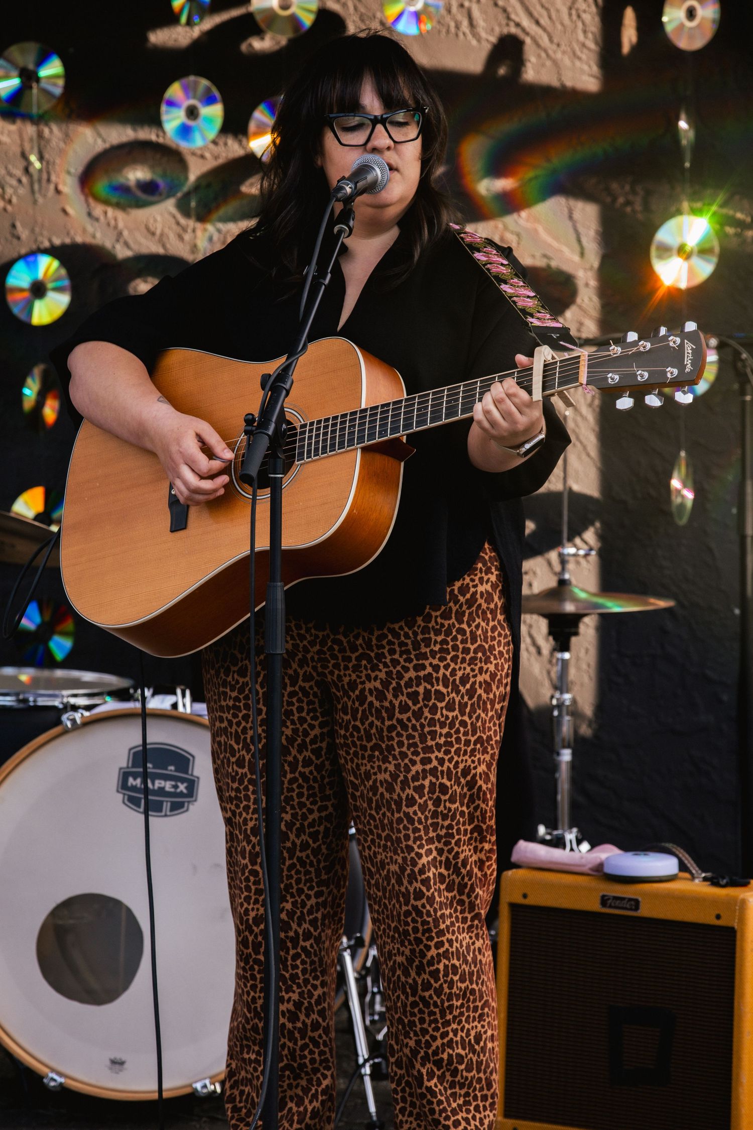Woman in leopard print pants playing acoustic guitar and singing with CDs decorating the background.