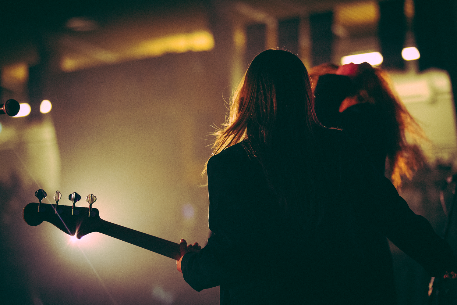 Two long-haired musicians on stage from behind, one holding bass guitar under warm concert lighting.