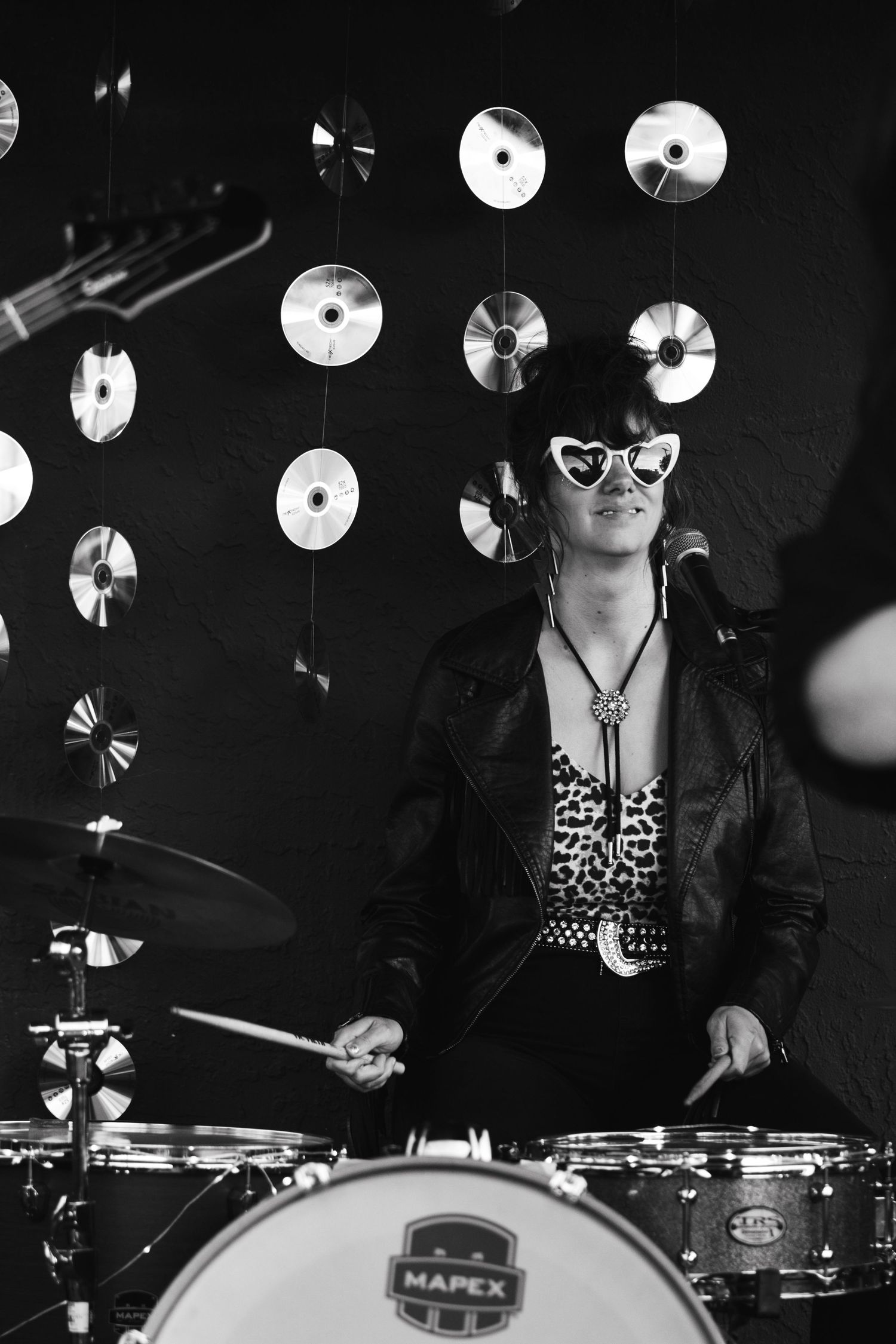Female drummer in leather jacket and heart sunglasses playing Mapex drums with CDs hanging in background.