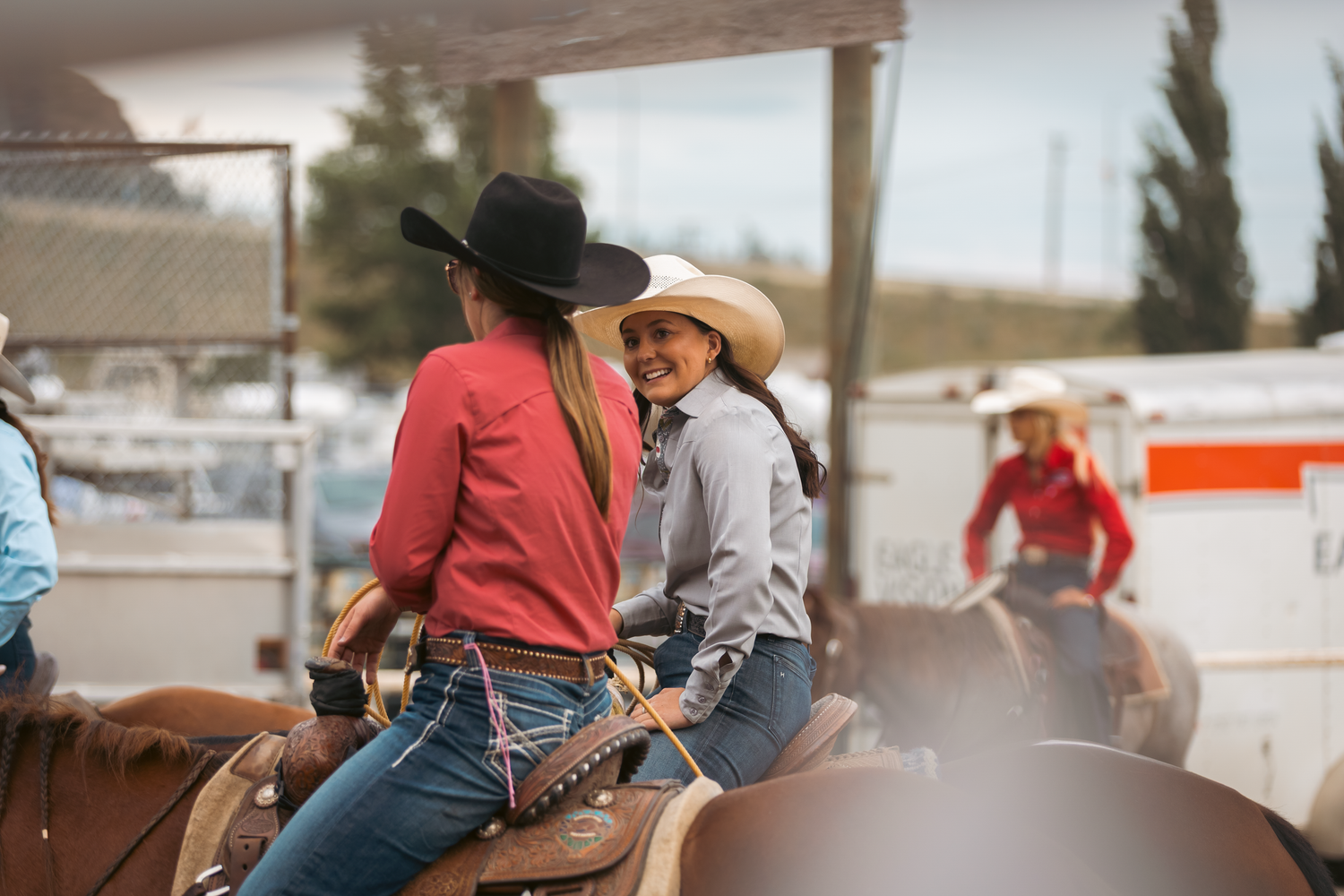Two smiling women in cowboy hats ride horses together at an outdoor rodeo event.