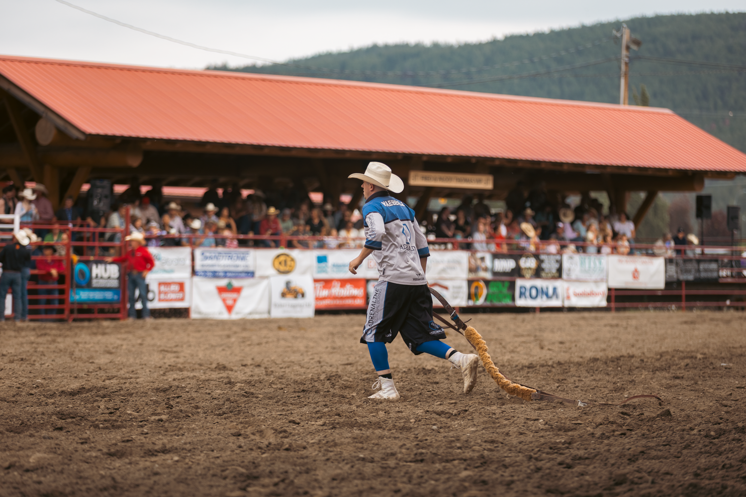 Rodeo clown in white hat and colorful outfit walks across a dirt arena carrying a rope at a rodeo.