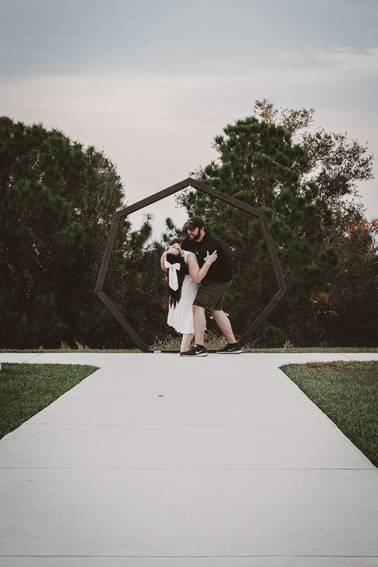 Couple embracing under a geometric hexagonal metal arch on a pathway surrounded by lush green trees at dusk.