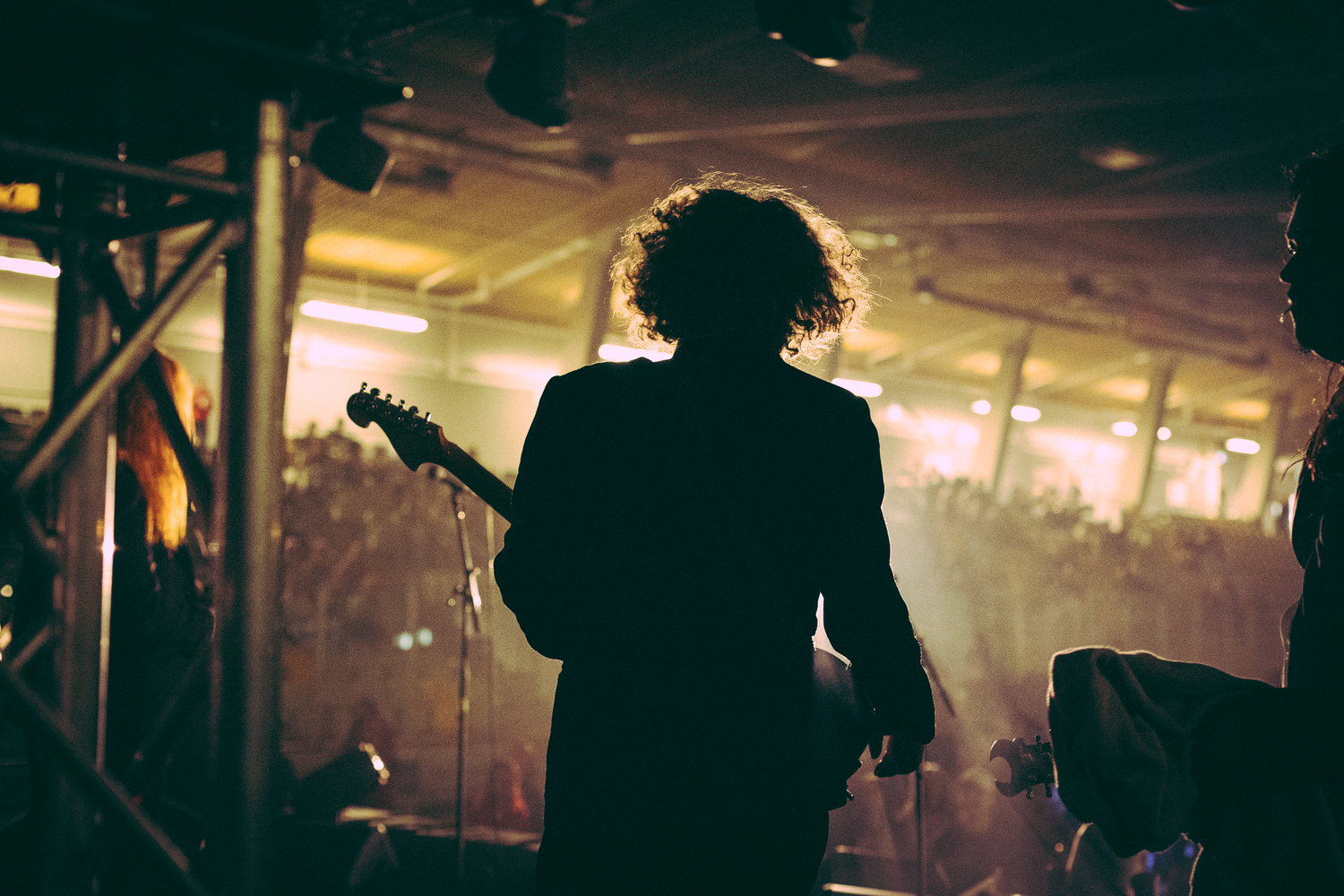 Silhouetted curly-haired guitarist standing on stage holding electric guitar before a glowing warm-lit concert crowd.