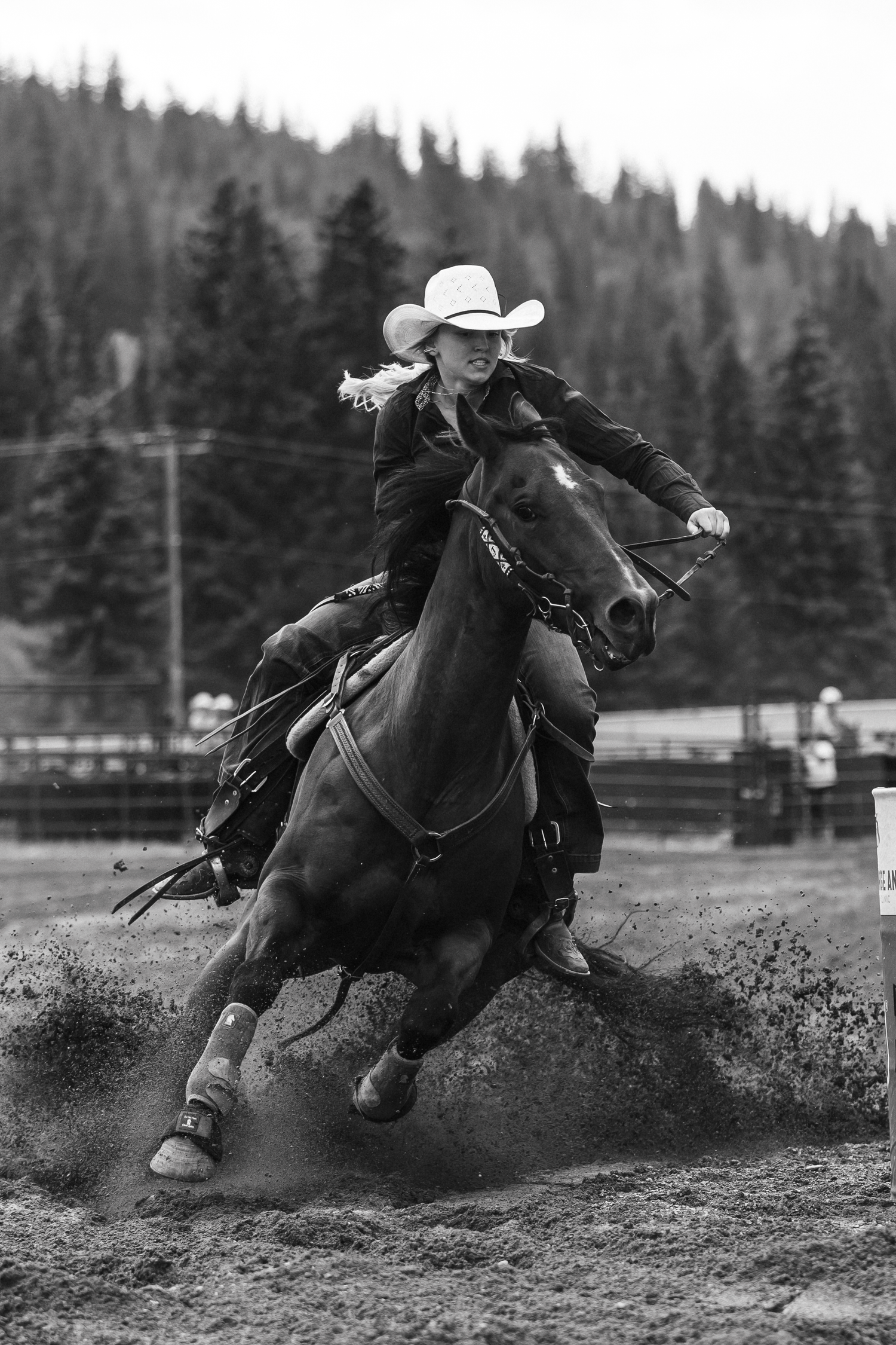 Black and white photo of a cowgirl in white hat riding horse at full gallop during barrel racing rodeo event.