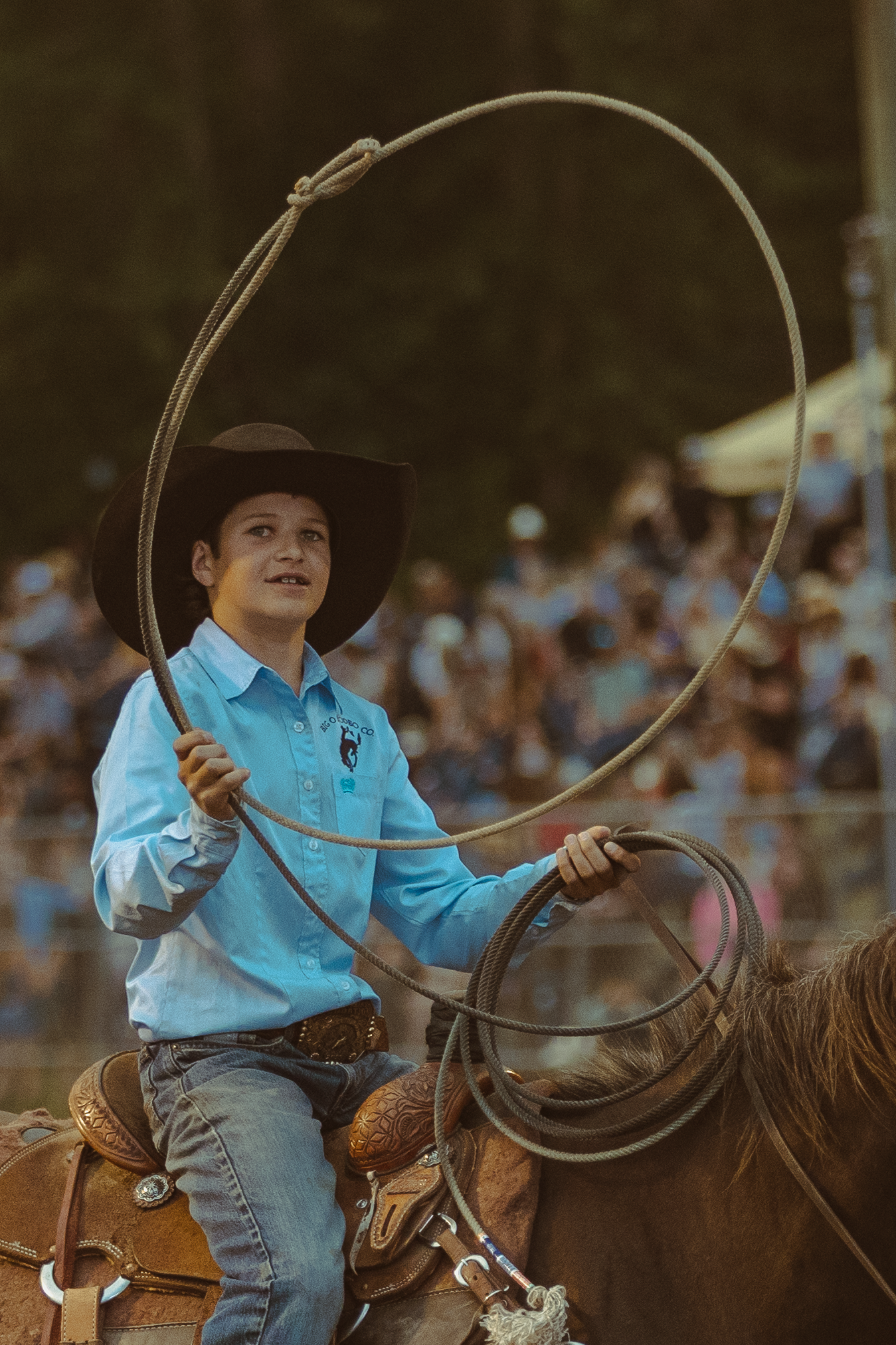 A young cowboy in a light blue shirt spins a lasso while riding a horse at a rodeo.