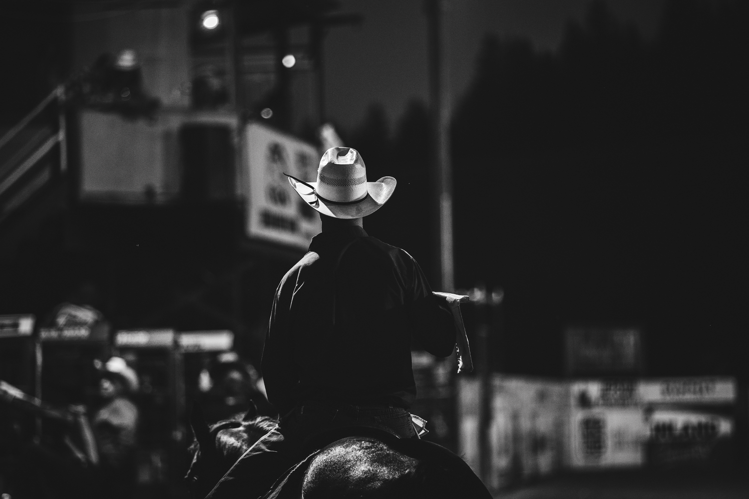 Silhouette of cowboy in hat sitting on horse at nighttime rodeo arena.