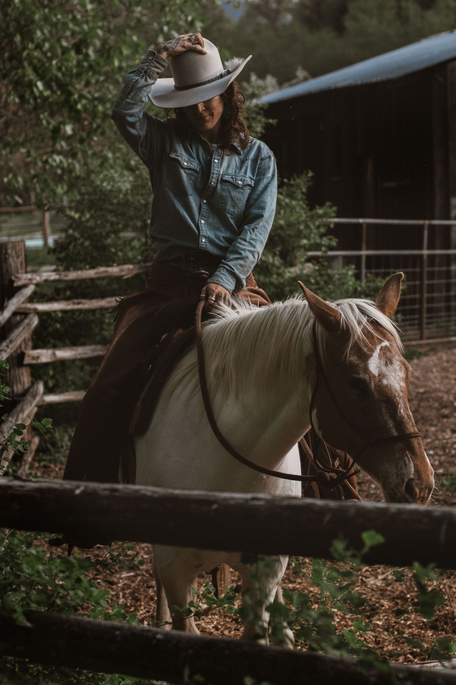 A woman in a denim shirt and cowboy hat rides a paint horse through a rustic wooden fence corral.
