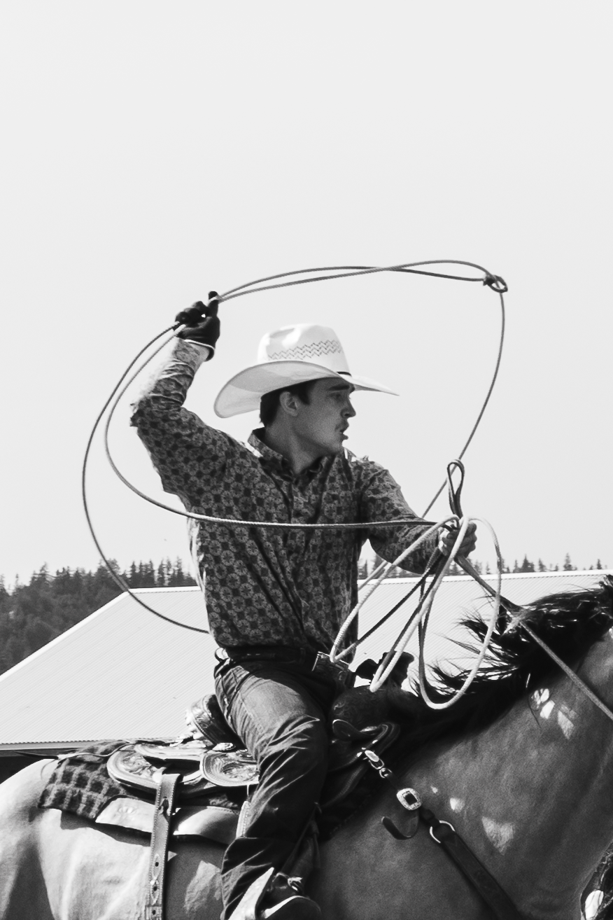Black and white photo of a cowboy on horseback swinging a lasso rope in an open field.