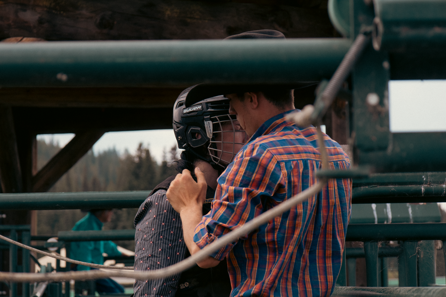 A man in a plaid shirt helps a rider fasten a protective helmet at a rodeo chute.
