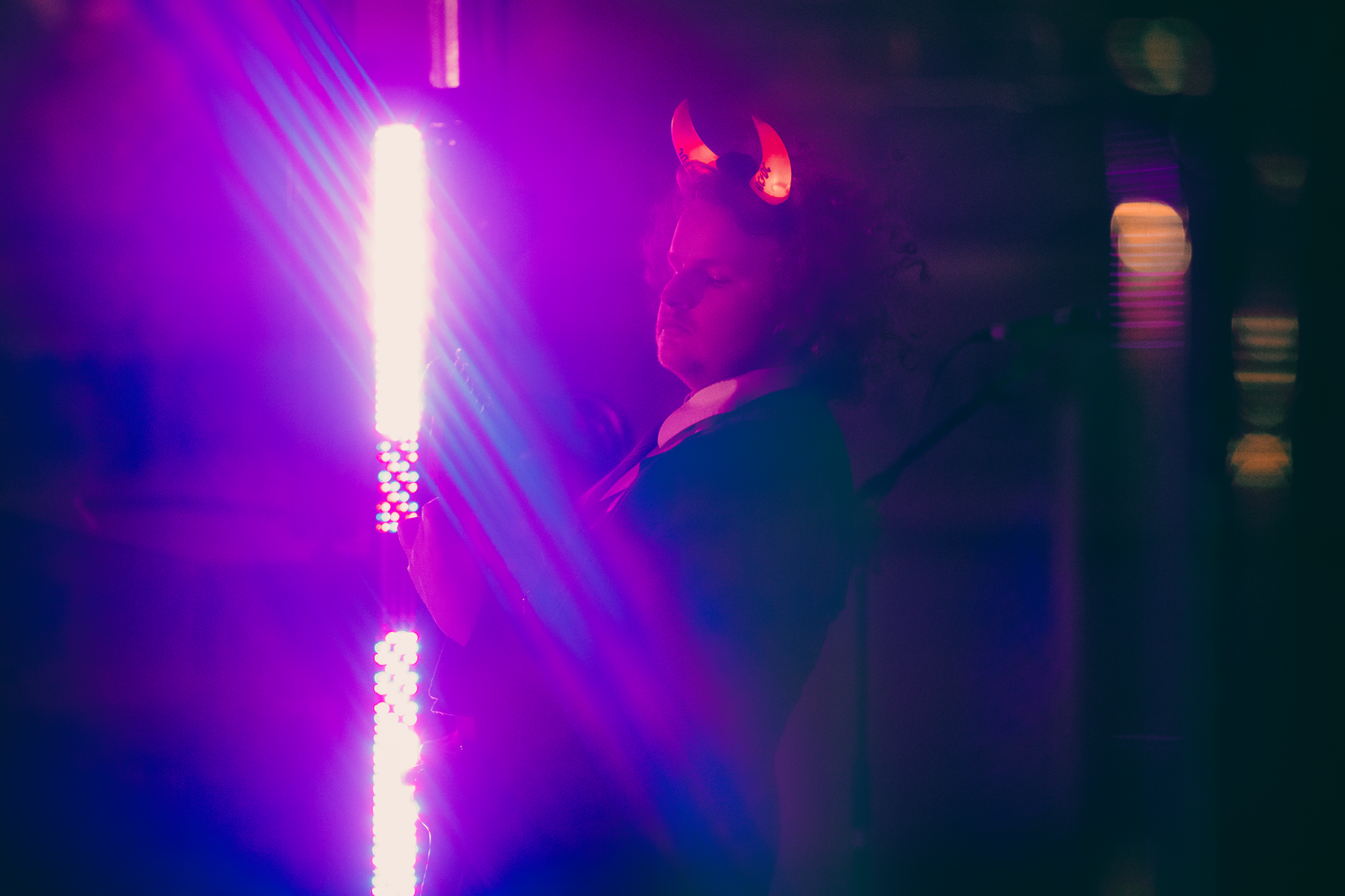 Performer wearing devil horns costume posing beside glowing neon light column at live music event.