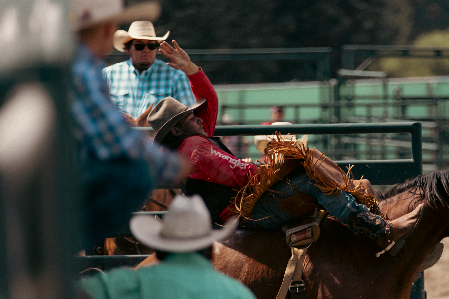 Rodeo rider in red shirt on bucking horse, with cowboys in white hats watching at a western rodeo event.