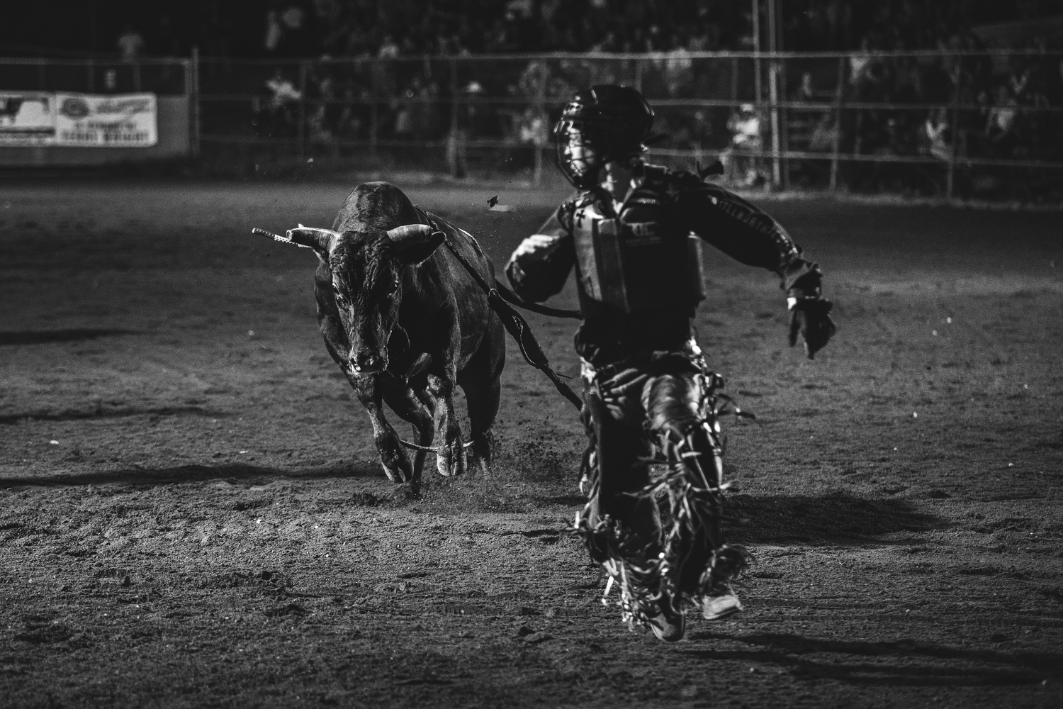 Black and white rodeo clown running from bull in dusty arena at night.