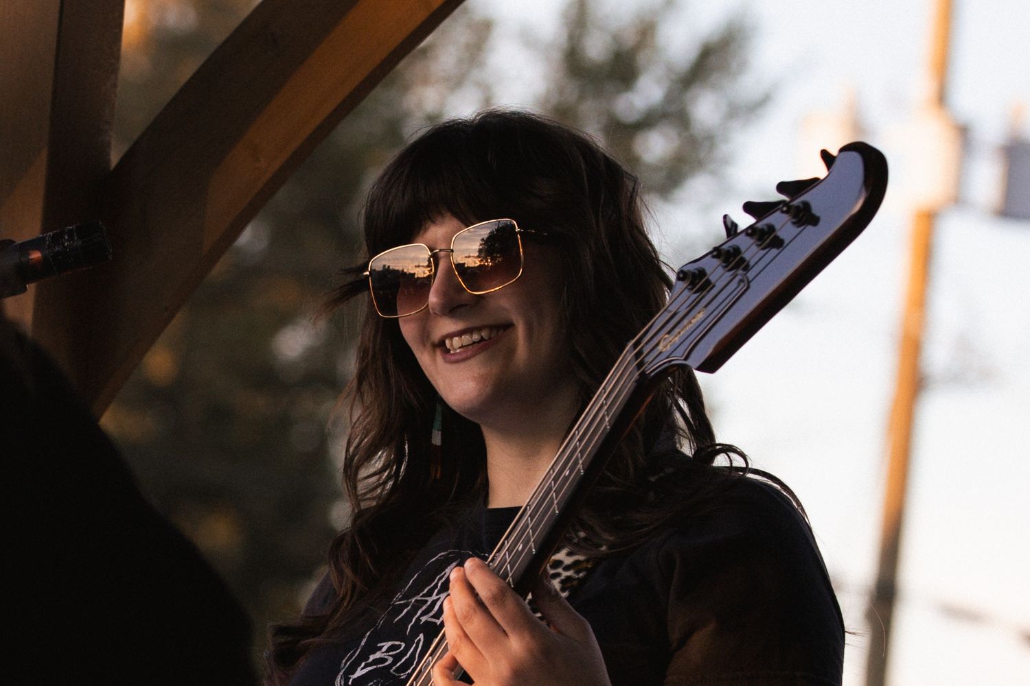 Smiling woman with sunglasses holding a bass guitar outdoors in bright sunlight.