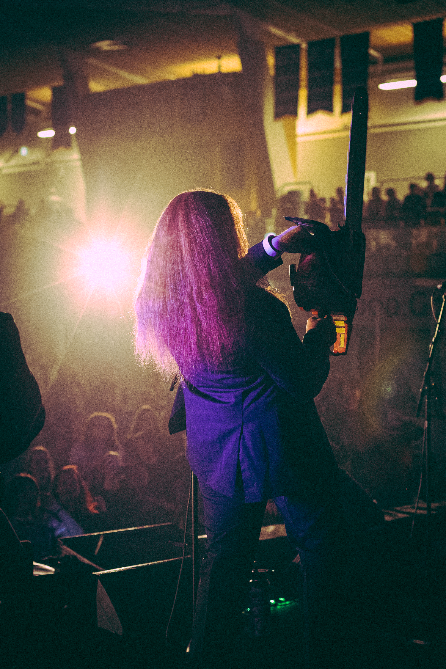 Long purple-haired performer on stage facing crowd, raising fist under bright golden concert spotlights.