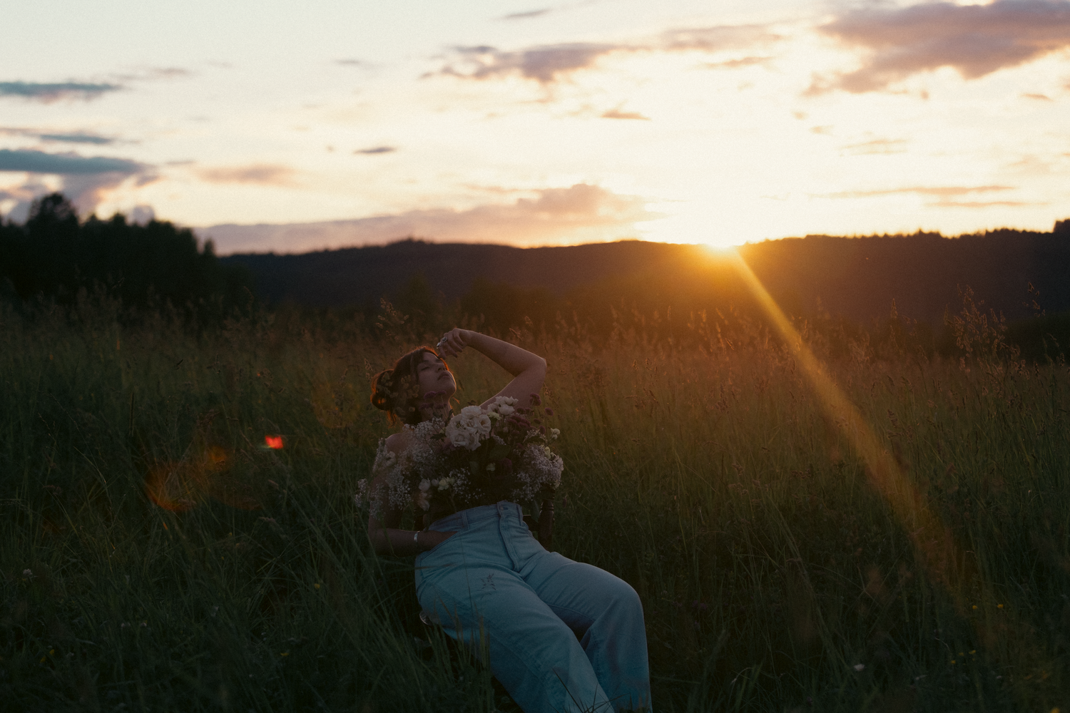 A woman sits in a golden field at sunset, basking in the warm glow of the setting sun over rolling hills.