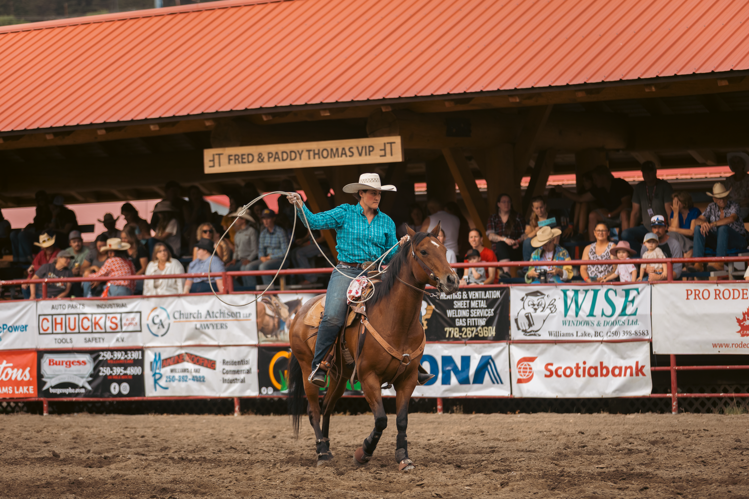 Female barrel racer in turquoise shirt rides a horse along the arena fence past sponsor signs at a rodeo.