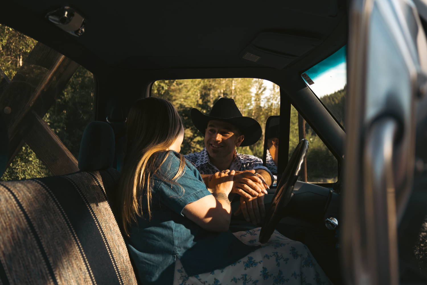 A bearded man in a cowboy hat plays guitar for a woman inside a sunlit vintage truck cab.