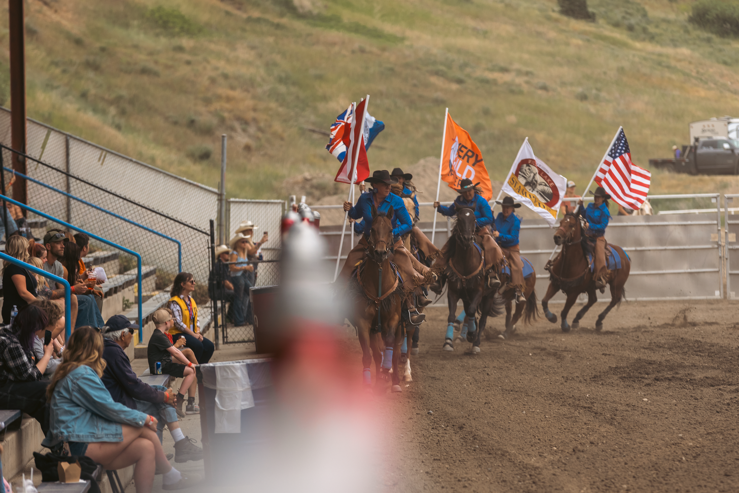 Multiple riders carrying colorful flags gallop past spectators in bleachers at an outdoor rodeo.