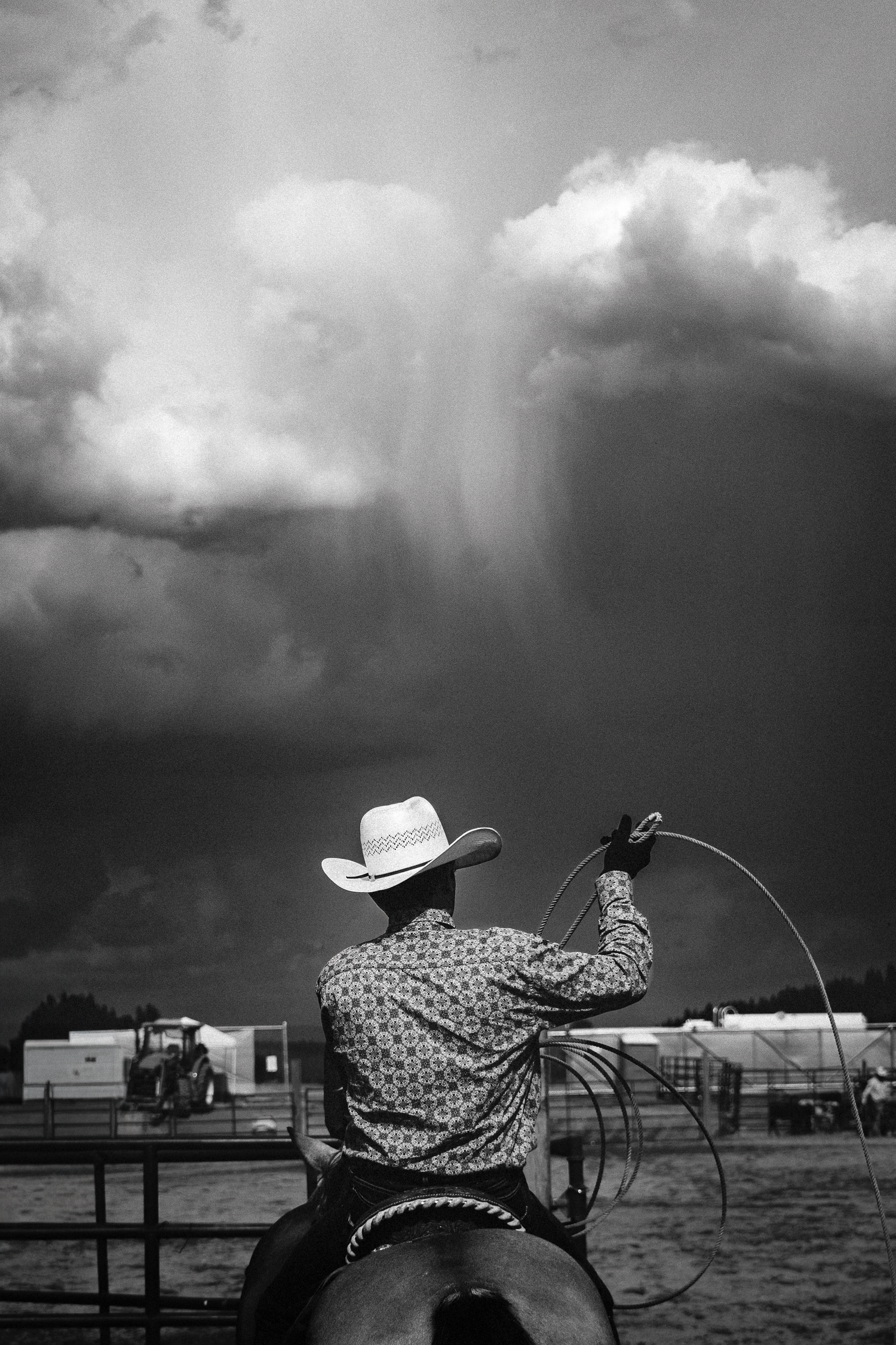 Black and white photo of a cowboy on horseback holding a lasso with dramatic storm clouds in the background.
