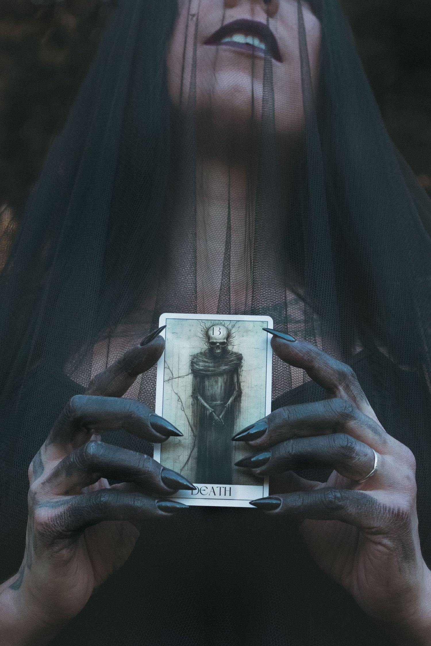 Veiled figure in black holding a Death tarot card with darkened hands and long black nails.